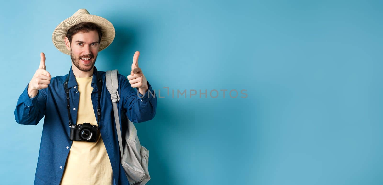 Handsome tourist going on summer vacation with camera and backpack pointing fingers at camera and inviting you, standing on blue background.