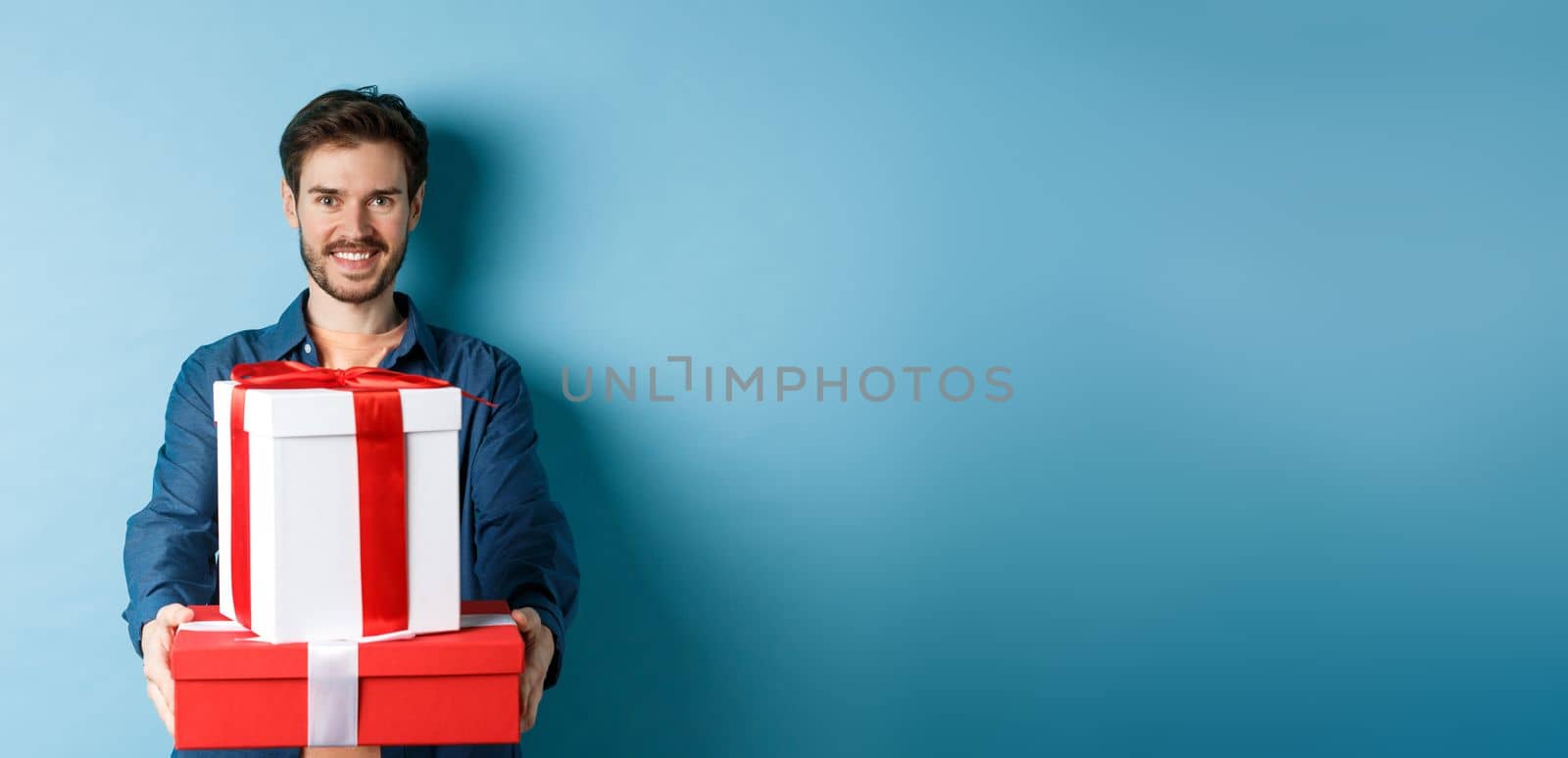 Happy valentines day. Handsome man giving girlfriend presents, holding gift boxes and smiling, standing over blue background.