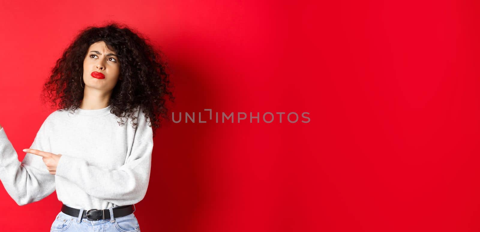 Skeptical frowning girl with curly hair, pointing and looking left hesitant, standing upset on red background by Benzoix