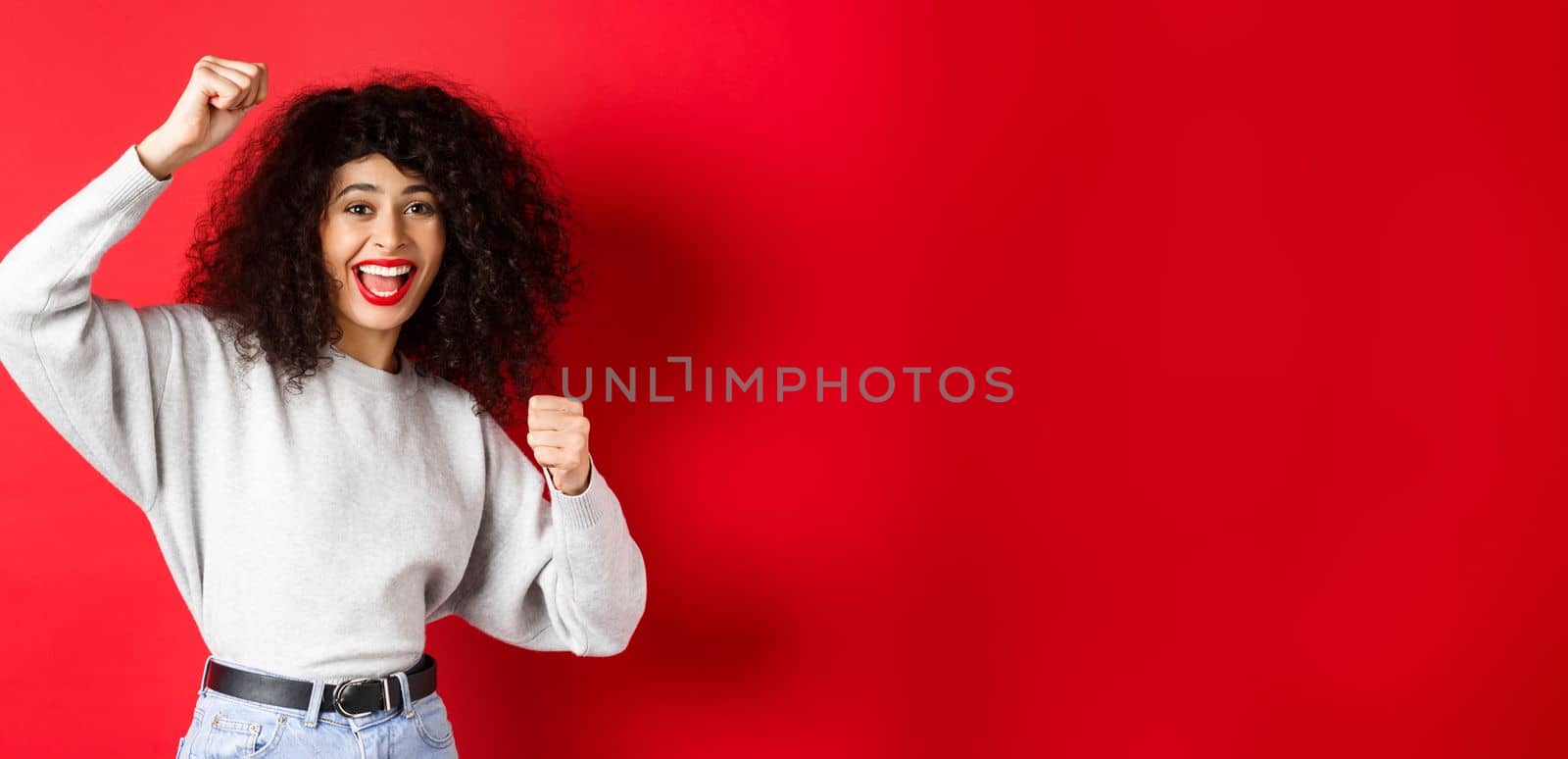 Cheerful young woman with curly hair, raising hand up and celebrating win, achieve goal or success, standing on red background.