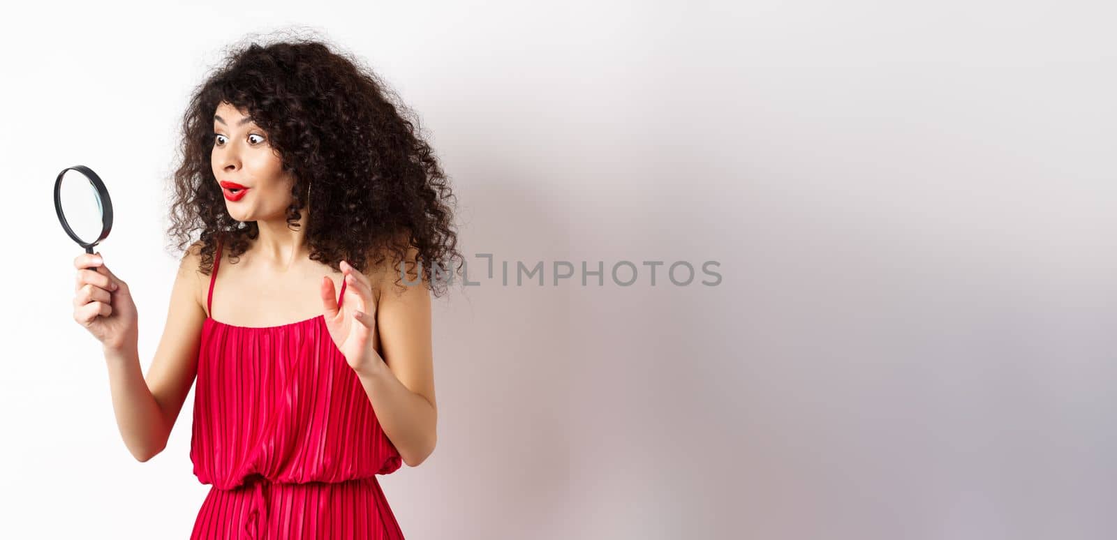 Excited woman in red dress look through magnifying glass and smiling, found interesting promo, standing on white background by Benzoix