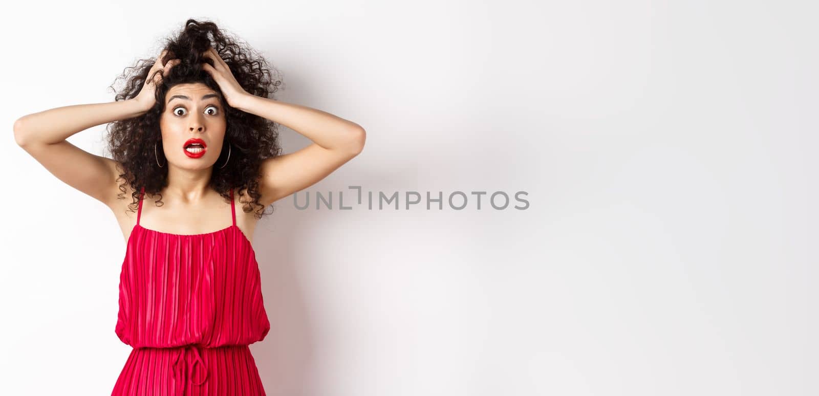 Shocked young woman holding hands on head and panicking, staring frustrated at camera, wearing red dress and makeup, white background by Benzoix