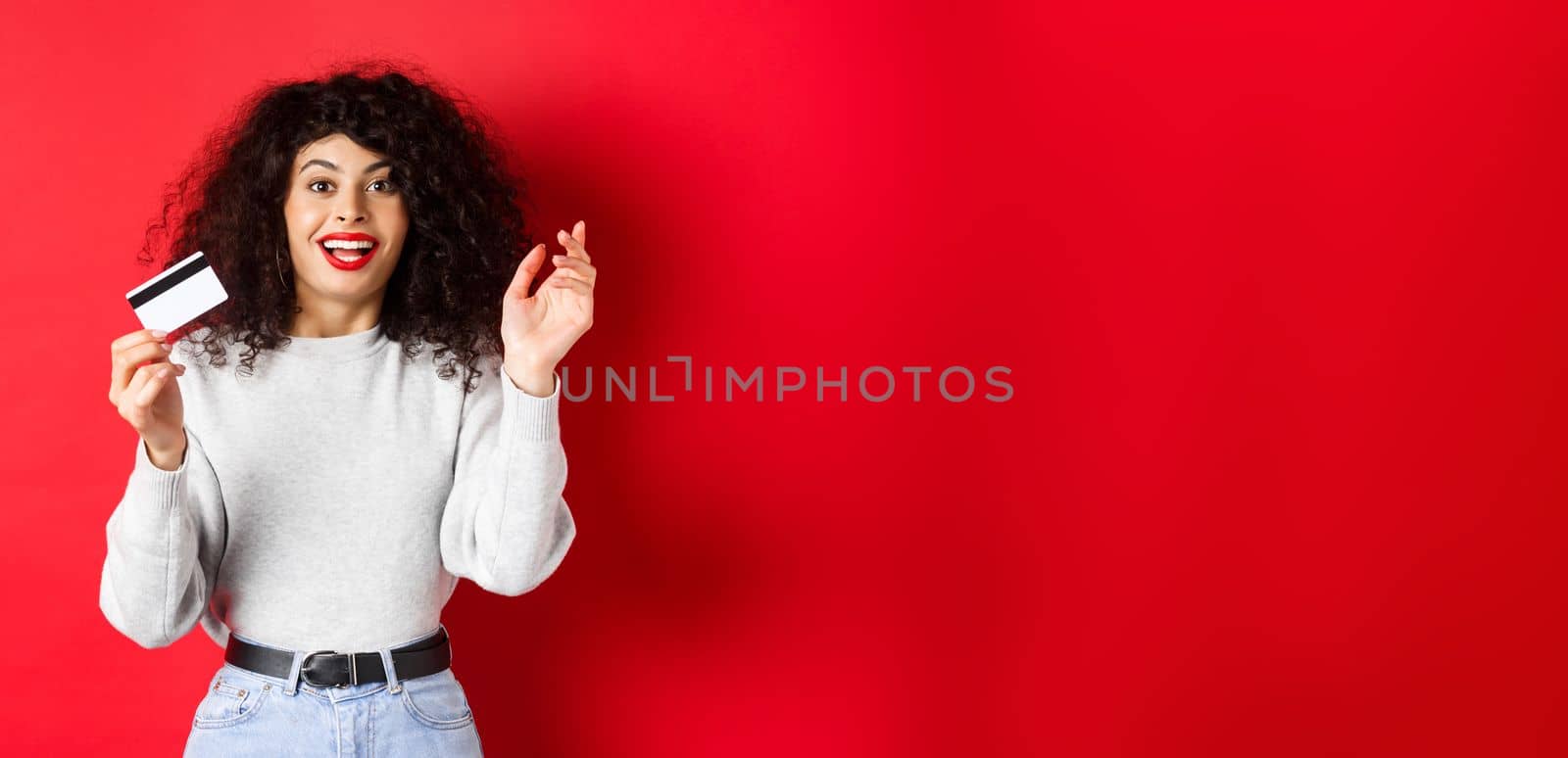 Excited woman telling about special deal, showing plastic card and looking happy, going shopping, standing on red background.