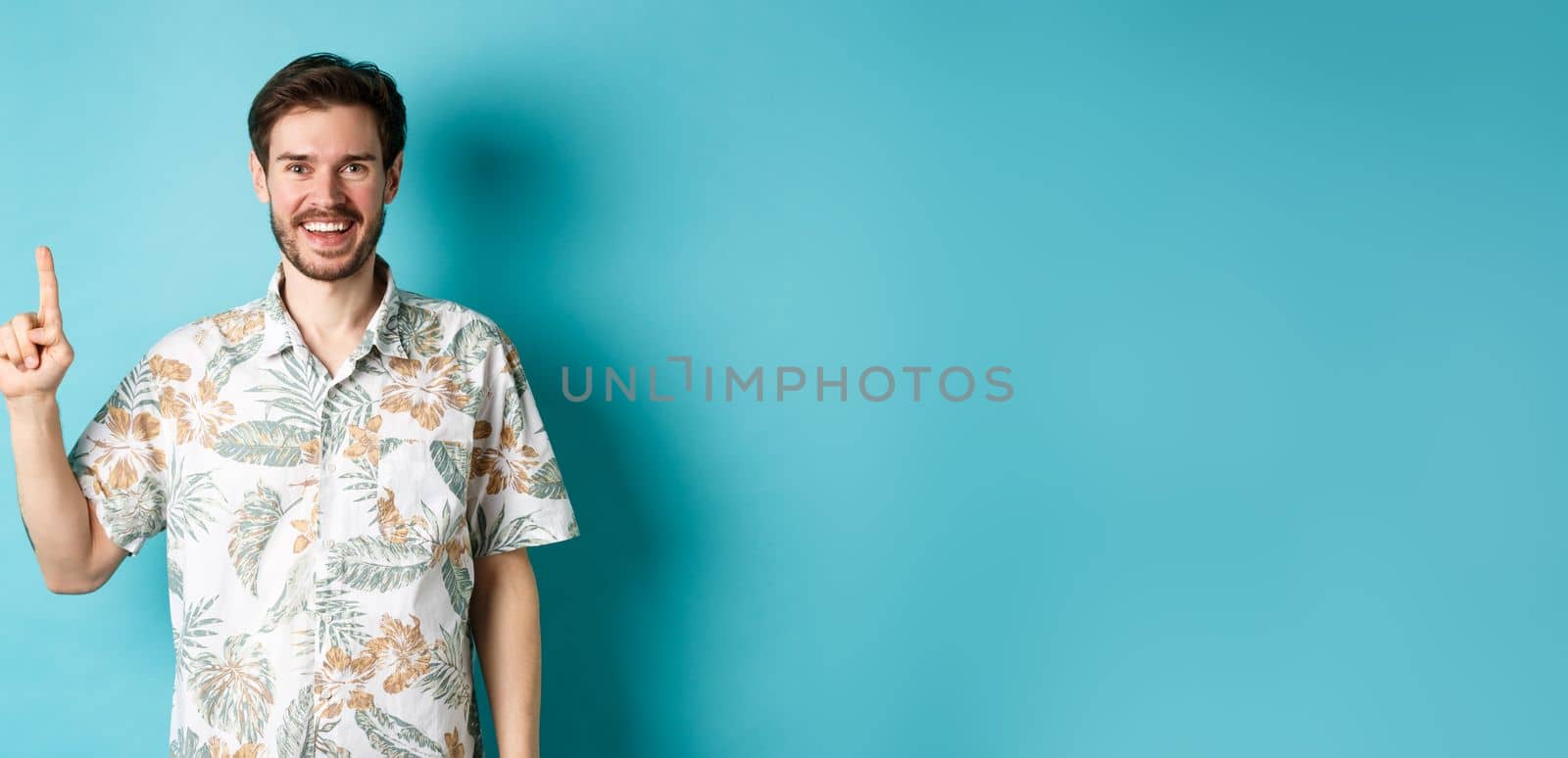 Happy tourist in hawaiian shirt smiling and pointing finger up, showing logo promotion, standing on blue background.