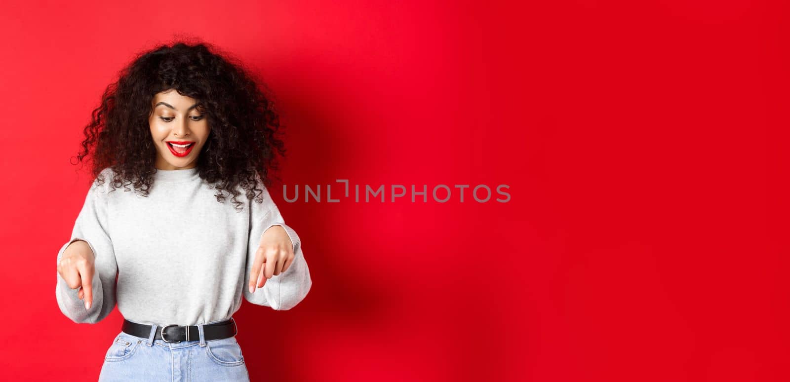 Dreamy beautiful woman with curly hair, pointing and looking down excited, checking out promo, standing against red background by Benzoix