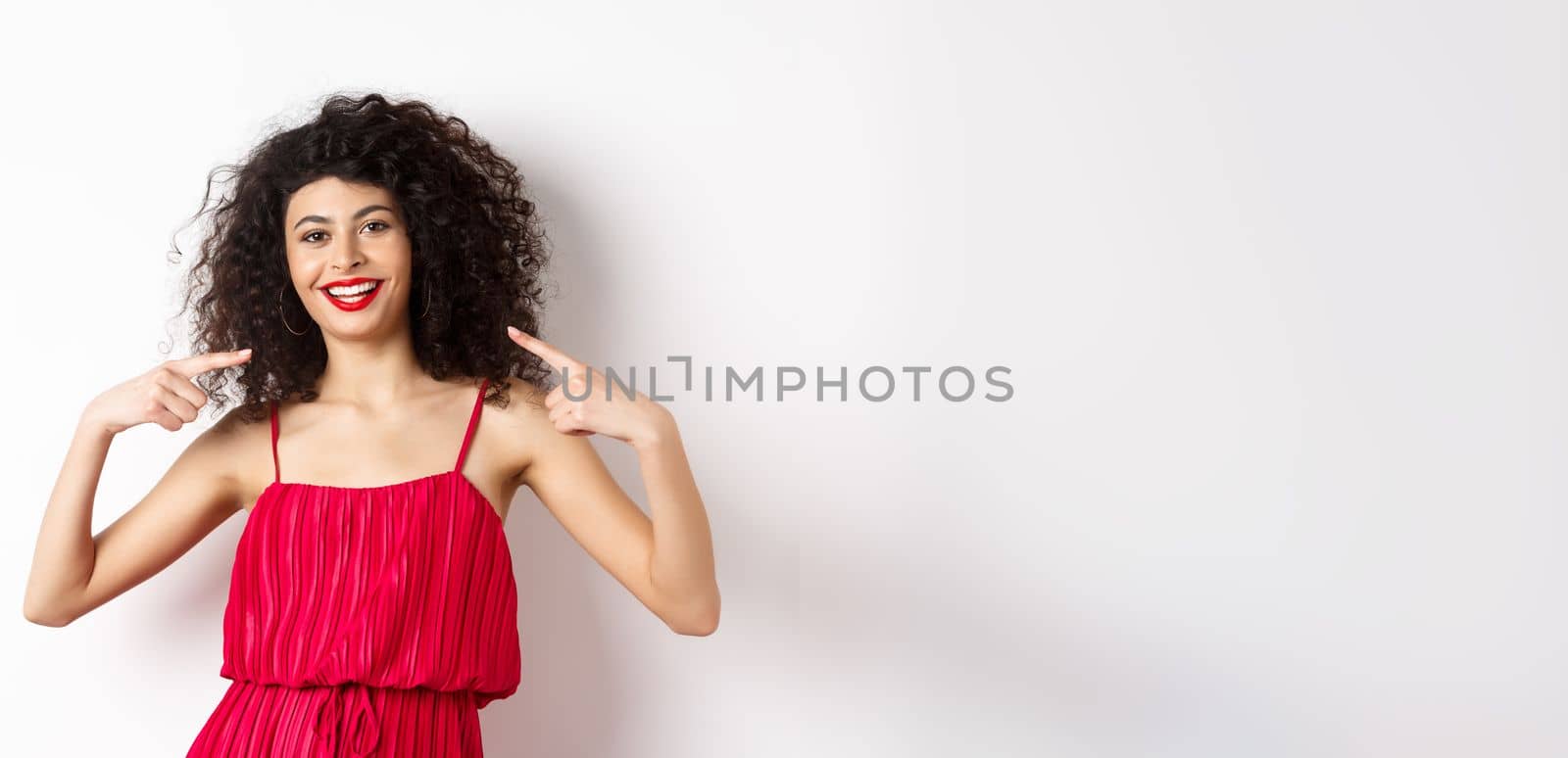 Beautiful woman with curly hairstyle, makeup and red dress, pointing at herself, smiling with white teeth, standing on white background by Benzoix