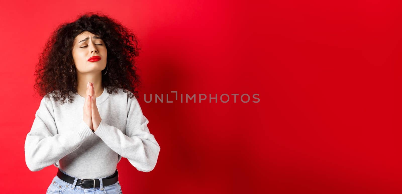 Desperate young woman pleading God, making wish and supplicating, holding hands in pray with eyes close and begging face, standing on red background.