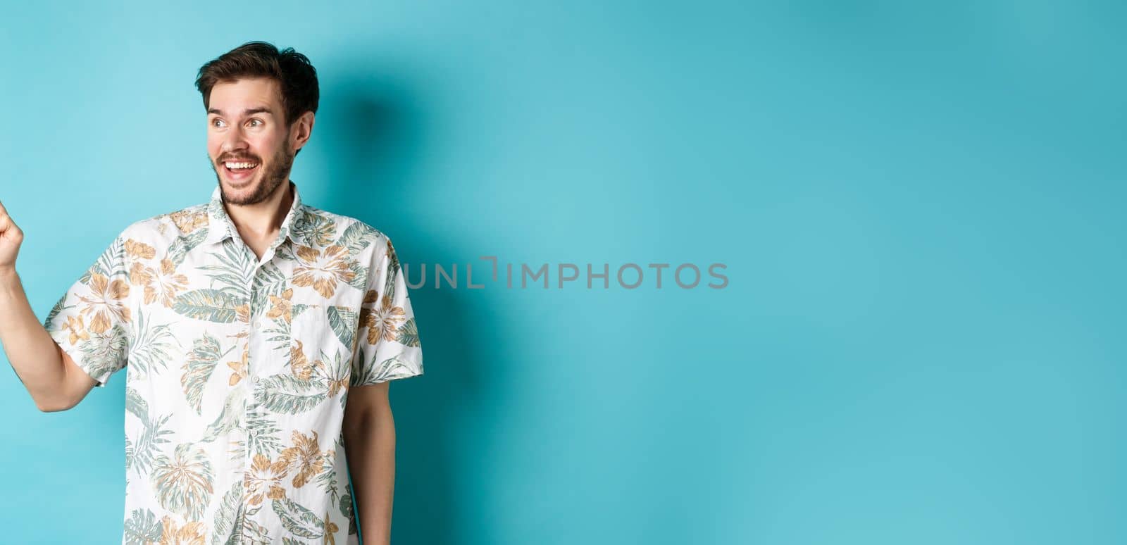 Excited smiling tourist pointing and looking aside at empty space, checking out promo offer, standing in hawaiian shirt on blue background.