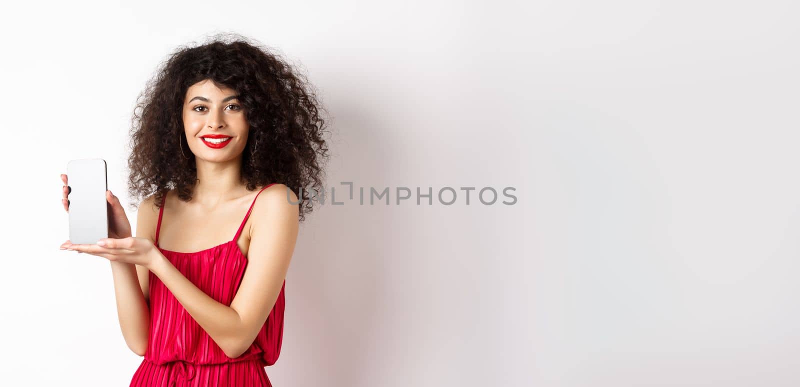 Elegant woman in red dress and makeup, showing blank smartphone screen and smiling, standing over white background. Copy space