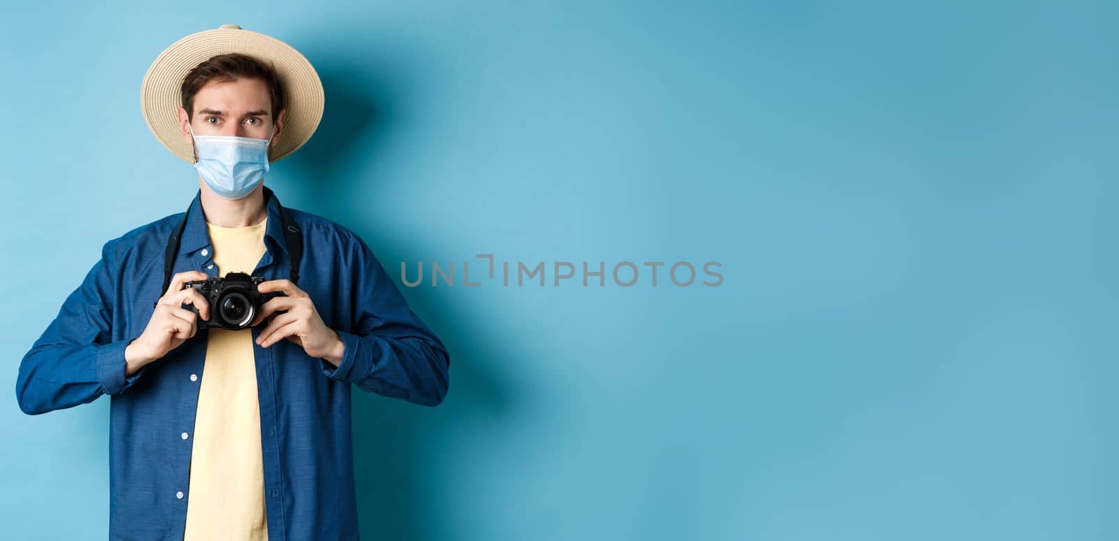 Covid-19, pandemic and travel concept. Cheerful tourist in summer hat and medical mask taking photos, holding camera, standing on blue background.