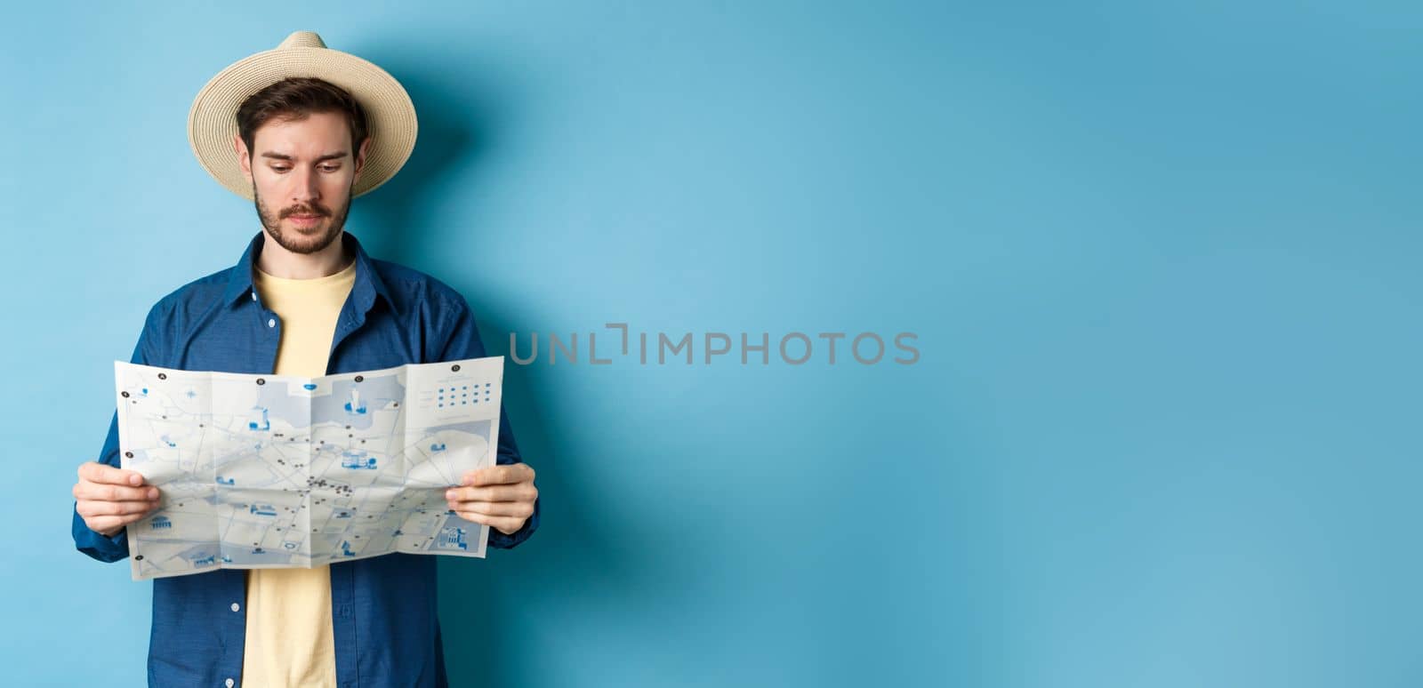 Handsome tourist with summer hat reading map, looking for sightseeing on vacation, standing on blue background by Benzoix