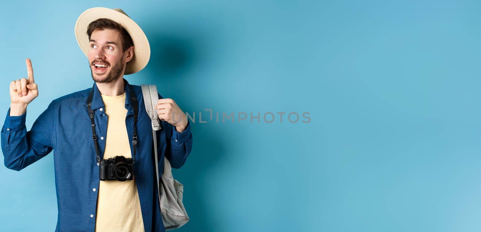 Handsome tourist looking and pointing up, travelling on summer vacation with backpack and camera, standing on blue background by Benzoix