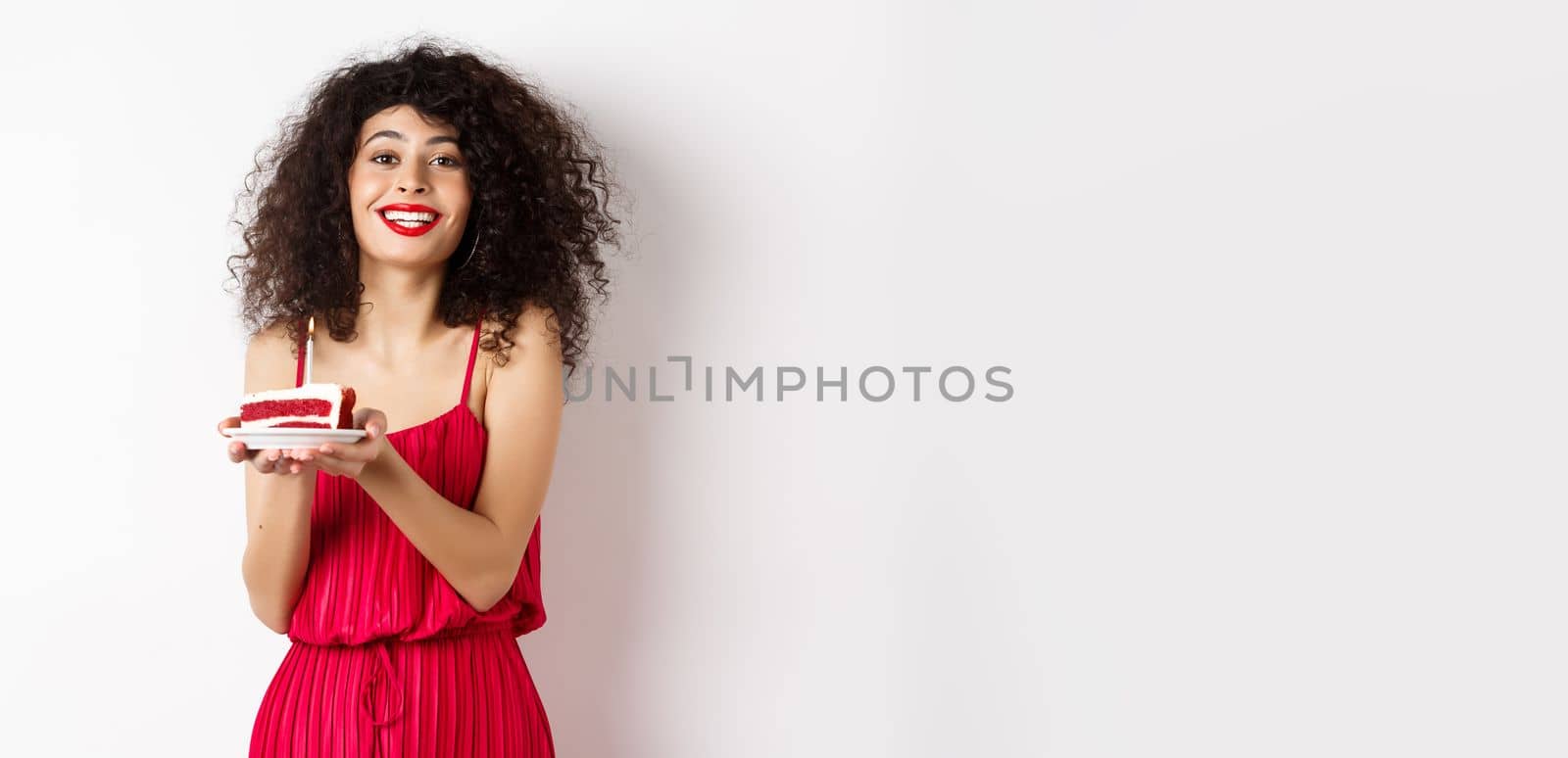 Beautiful lady in red dress celebrating birthday, holding piece of cake with candle and smiling, standing happy on white background.