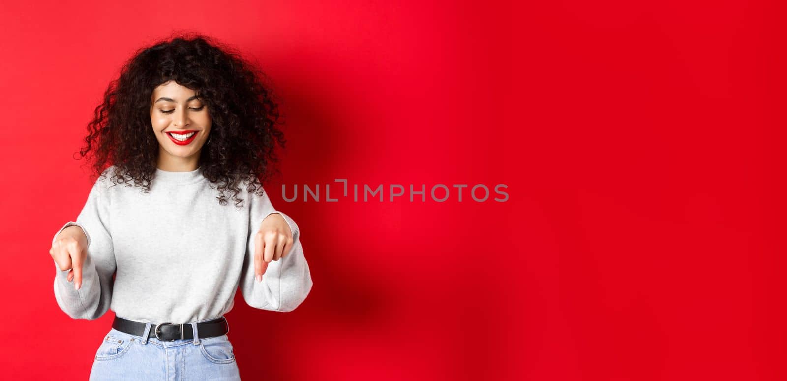 Modern woman with curly hairstyle and red lips, pointing fingers down and smiling happy, showing advertisement on empty space, studio background.