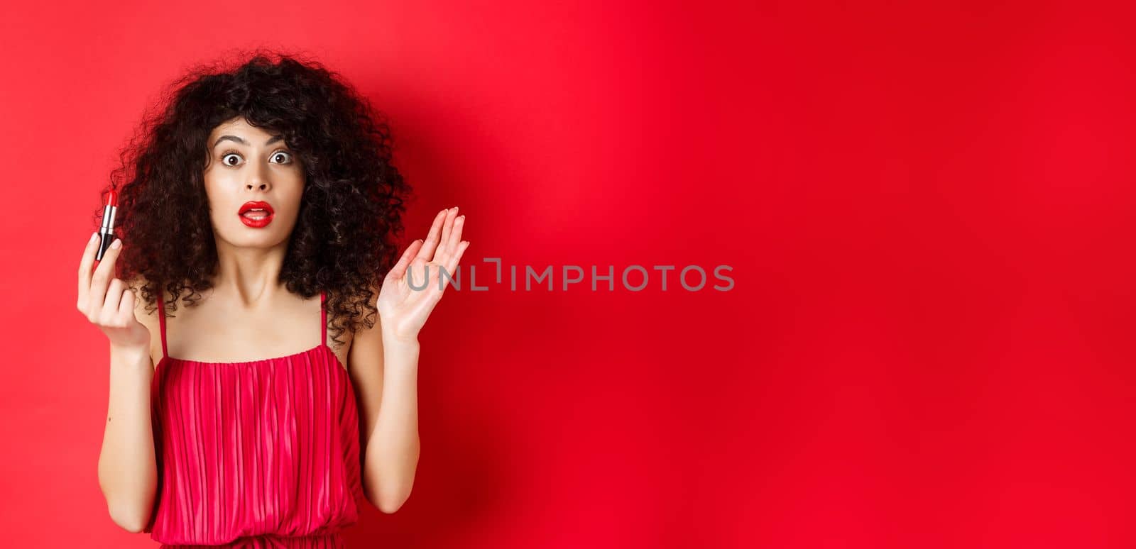 Beauty and makeup concept. Excited woman with curly hair, gasping as looking at camera, holding red lipstick, standing in dress on white background.