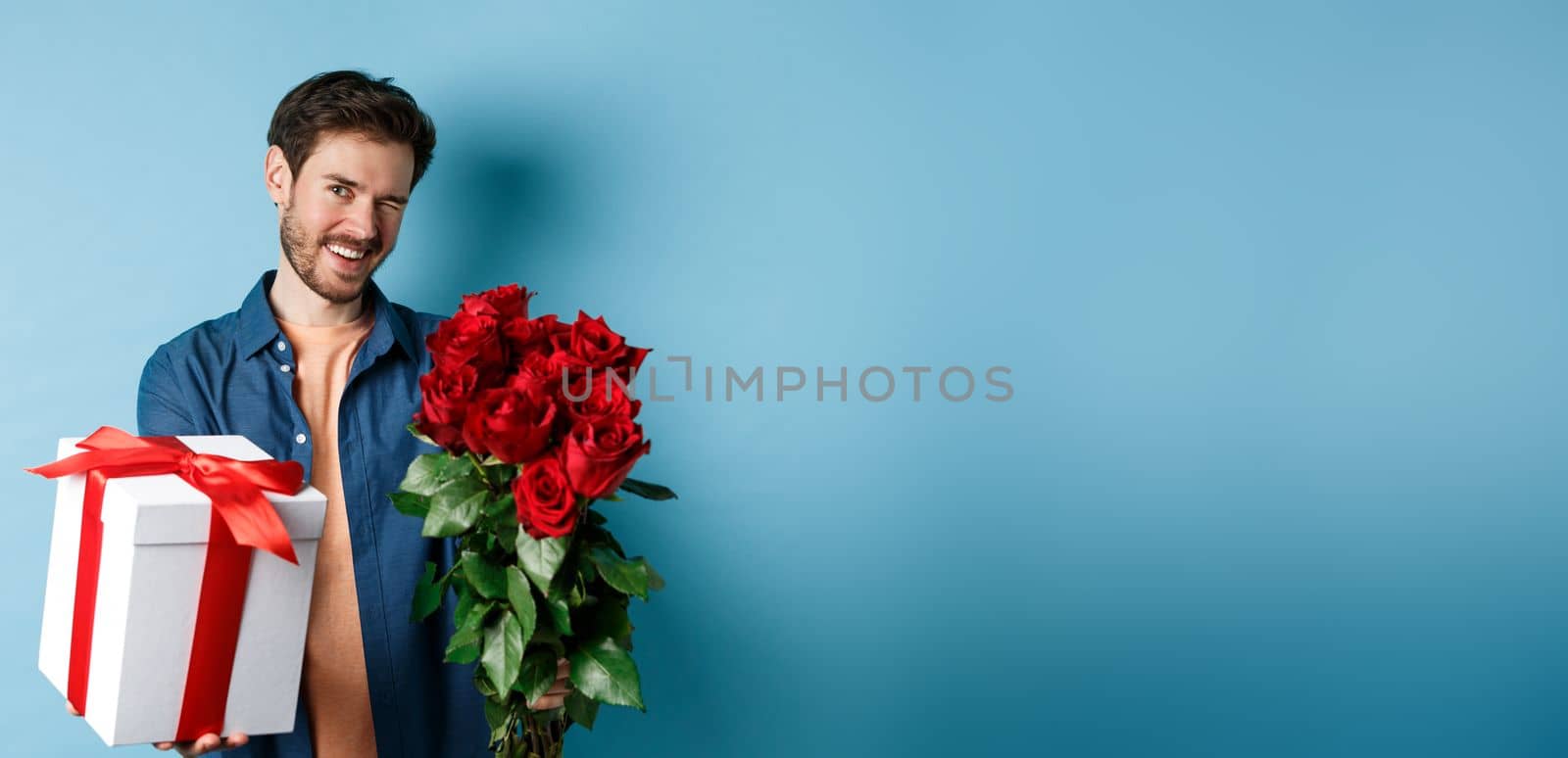 Love and Valentines day concept. Happy guy winking and smiling at camera, presenting a gift and bouquet of flowers to girlfriend, standing over blue background.