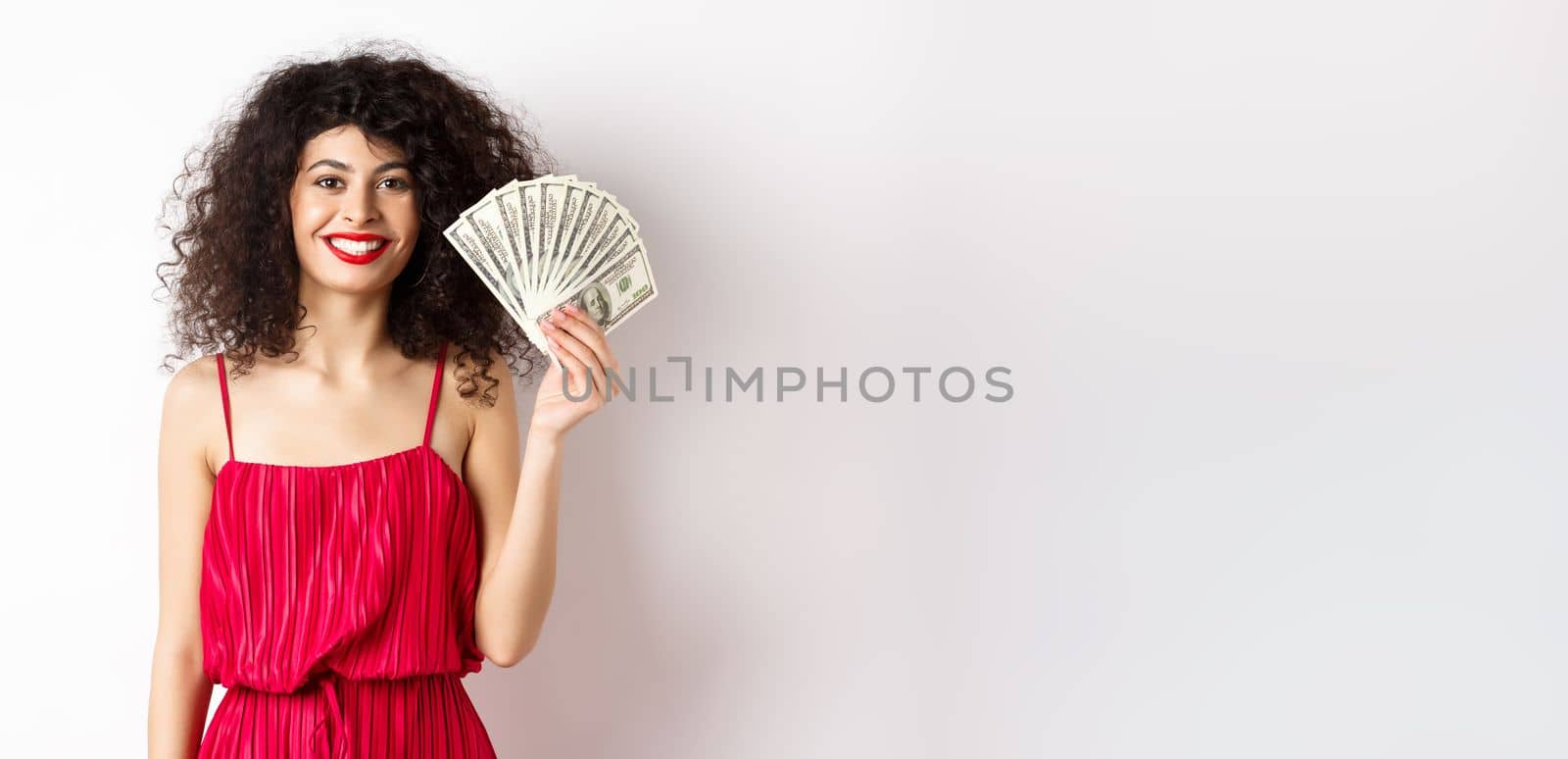 Stylish smiling woman in red dress showing money, standing on white background.