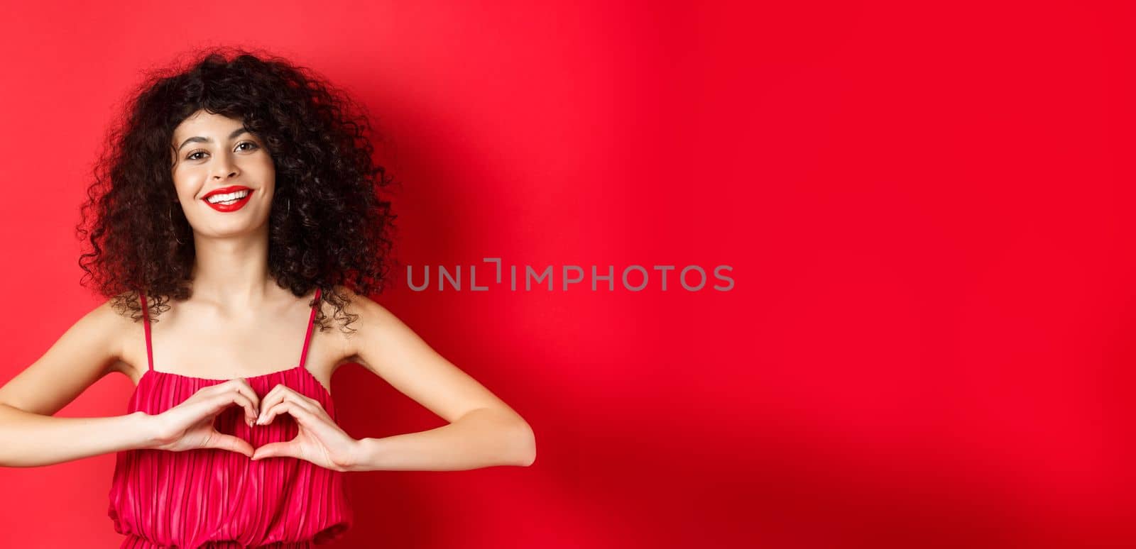 Lovers day. Beautiful woman celebrating valentines, showing heart sign and smiling, standing in romantic red dress on studio background.