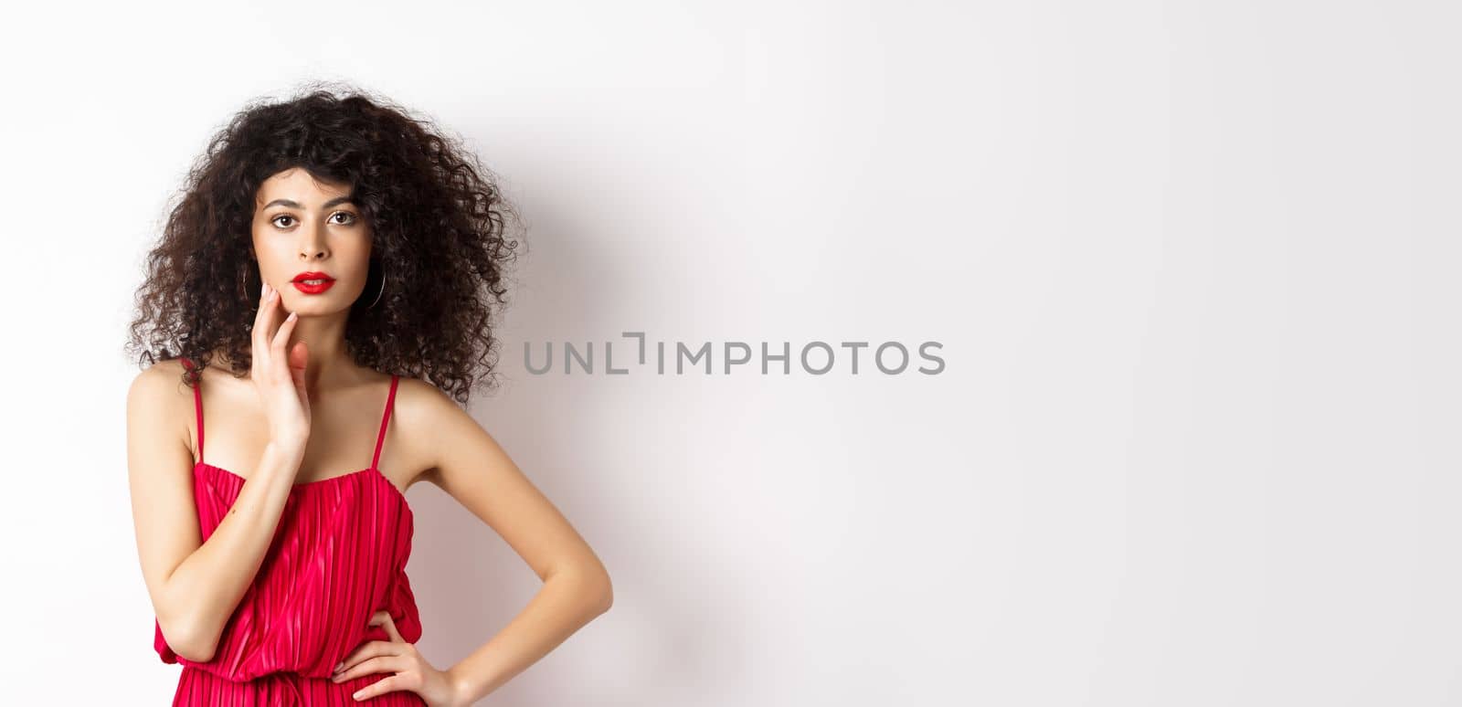 Beauty and fashion. Sensual female model with curly hair and red lips, wearing elegant dress, touching face and looking seductive, white background.