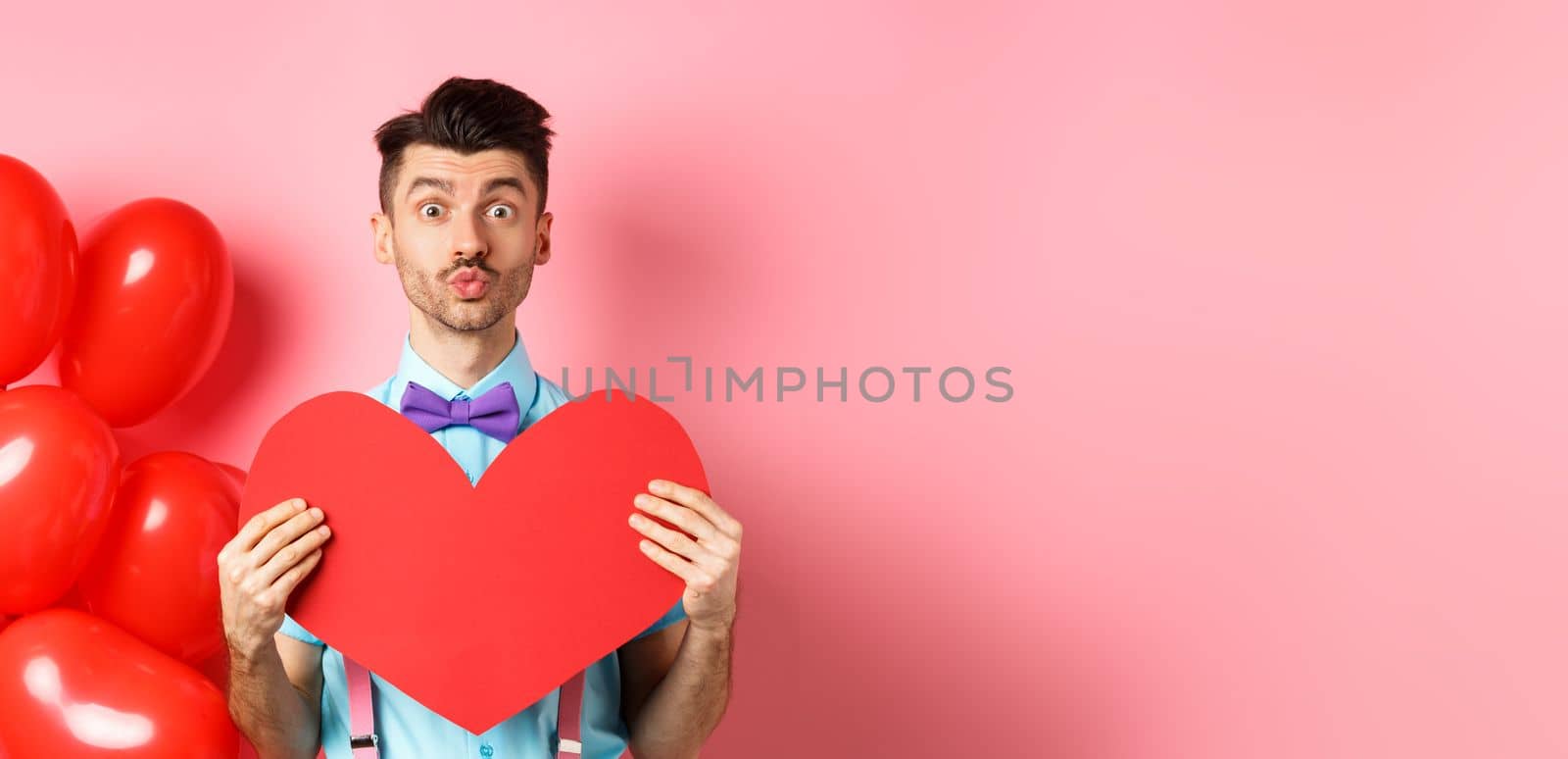 Valentines day concept. Cute guy pucker lips for kiss and showing romantic heart cutout, falling in love, standing over pink background.