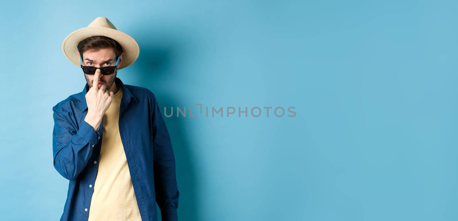 Handsome and cheeky guy put on sunglasses and straw hat, standing ready for summer vacation or tropical holiday, blue background.