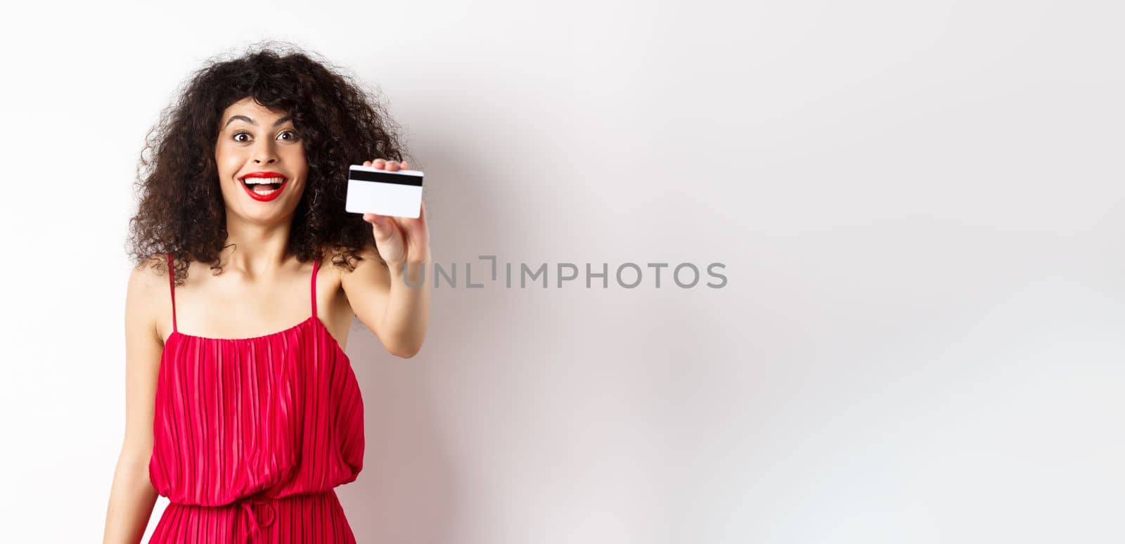 Shopping. Excited stylish lady with makeup, red dress, showing plastic credit card and smiling amazed, standing against white background by Benzoix