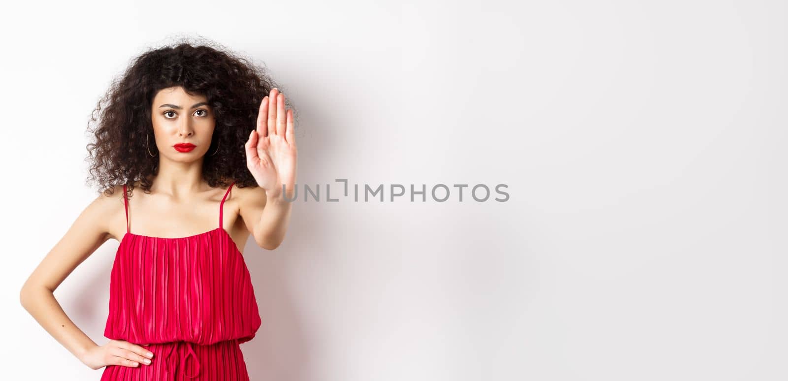 Serious and confident woman in red dress and makeup stretch out hand, tell to stop, prohibit and forbid something, standing over white background.