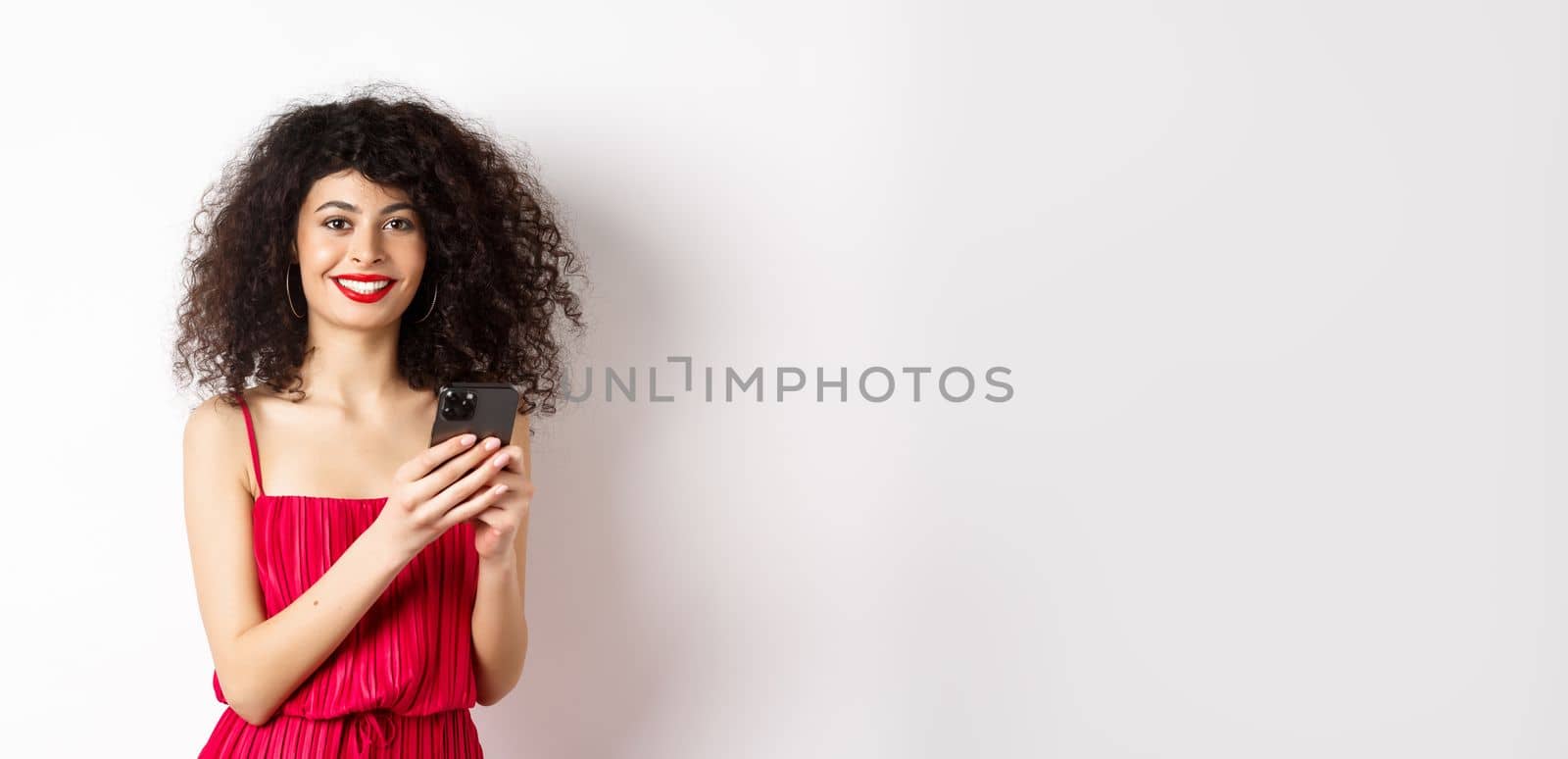 Cheerful woman with curly hair, using smartphone in red dress, smiling at camera, standing over white background by Benzoix