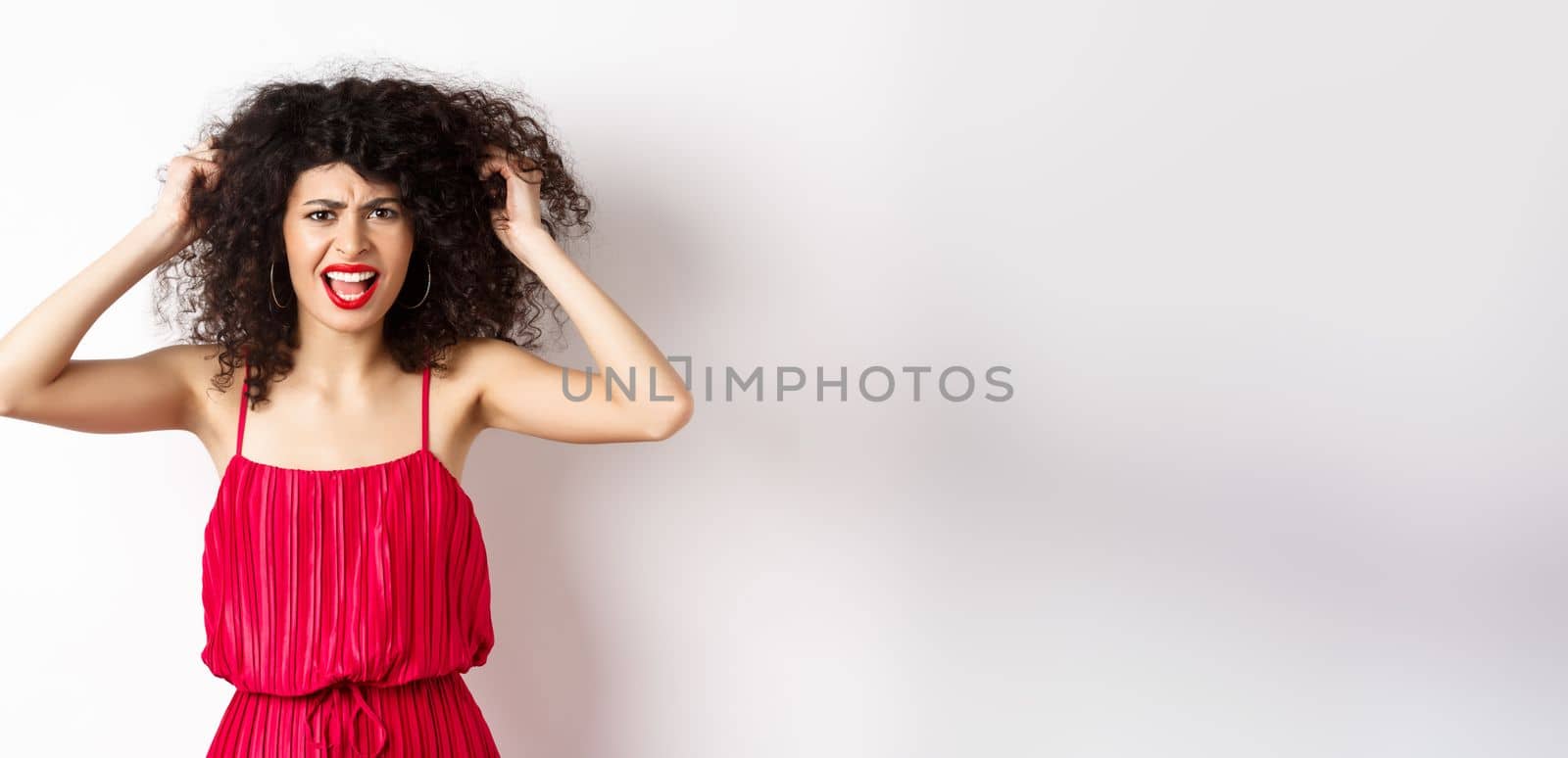 Frustrated curly woman in red dress, frowning and screaming angry, pull out hair and shout at camera, standing on white background.