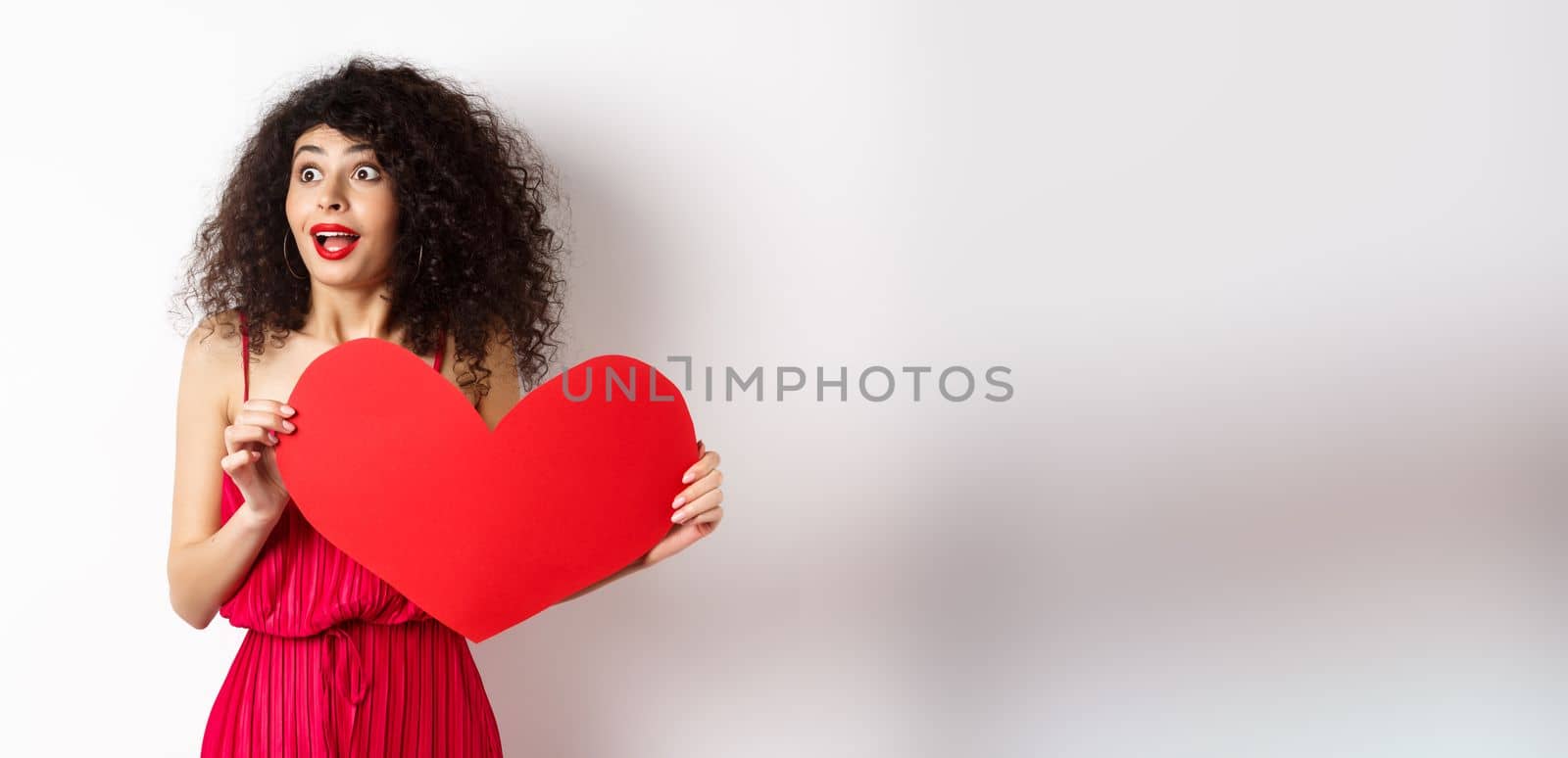 Valentines day. Young woman looking surprised left, seeing something amazing on lovers day, found true love, holding big red heart on chest, white background by Benzoix