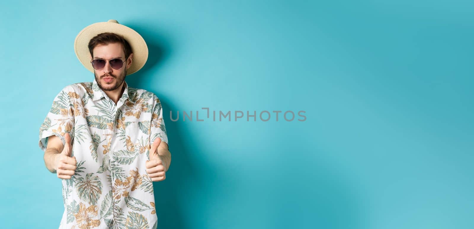 Handsome tourist enjoying summer holiday, wearing sunglasses and hawaiian shirt, showing thumbs-up in approval, standing on blue background.