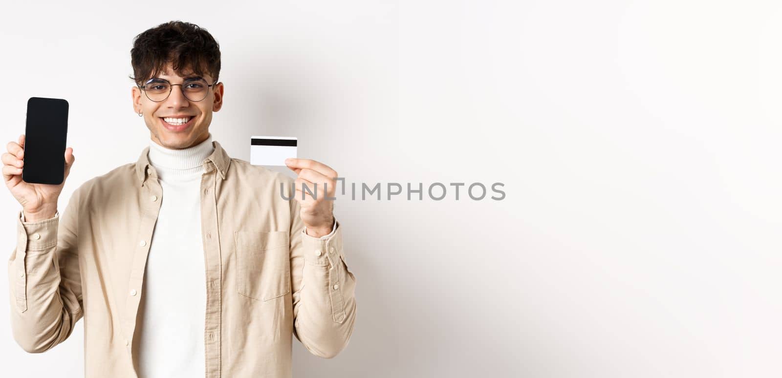 Online shopping. Natural guy in glasses showing empty smartphone screen and plastic credit card, smiling pleased, recommending bank, standing on white background.