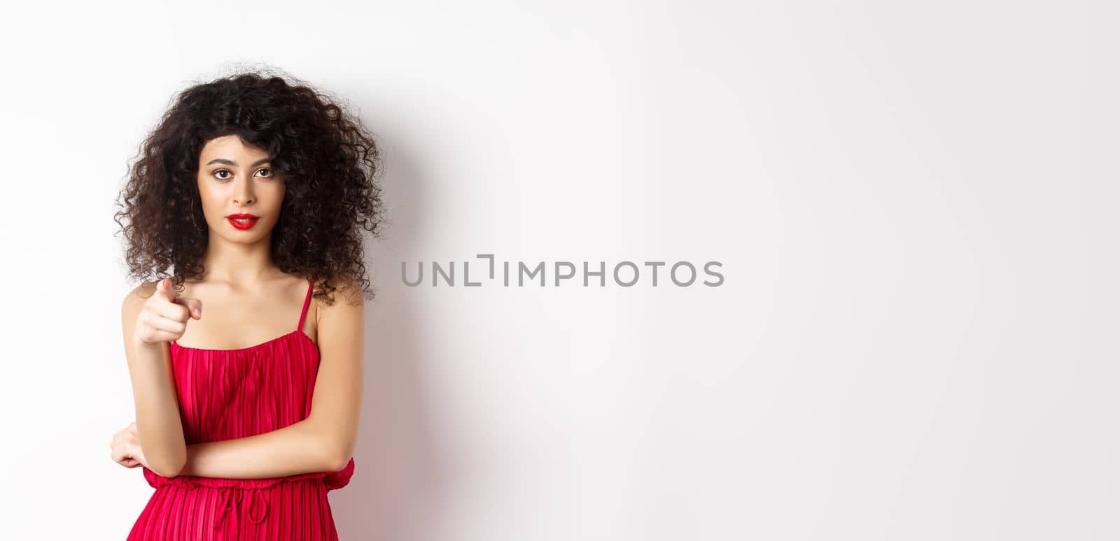 Confident young woman in red dress and makeup, pointing at camera, choosing you, inviting to event, standing over white background.
