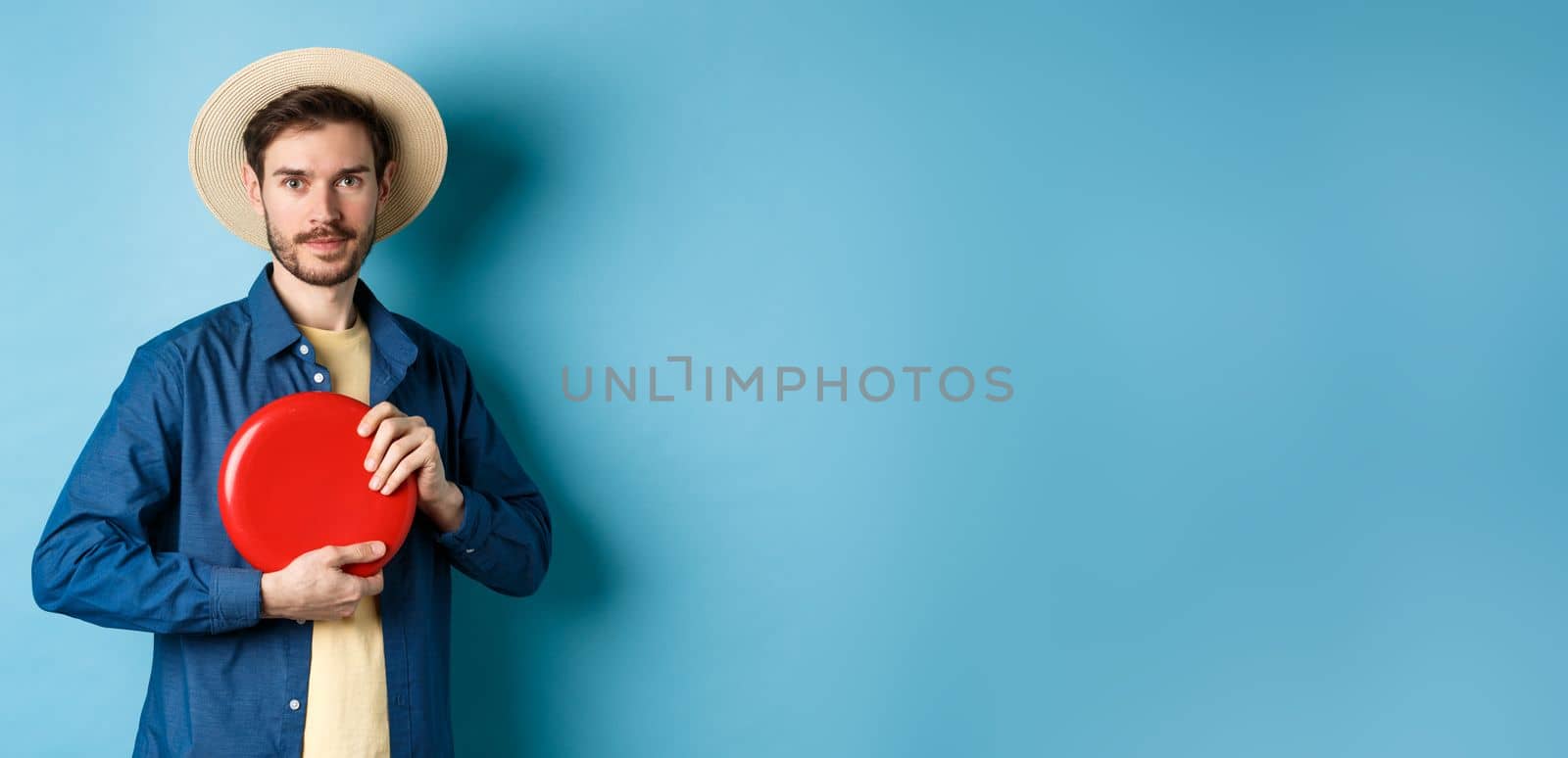 Young man throwing frisbee, playing on summer vacation, standing in straw hat on blue background by Benzoix