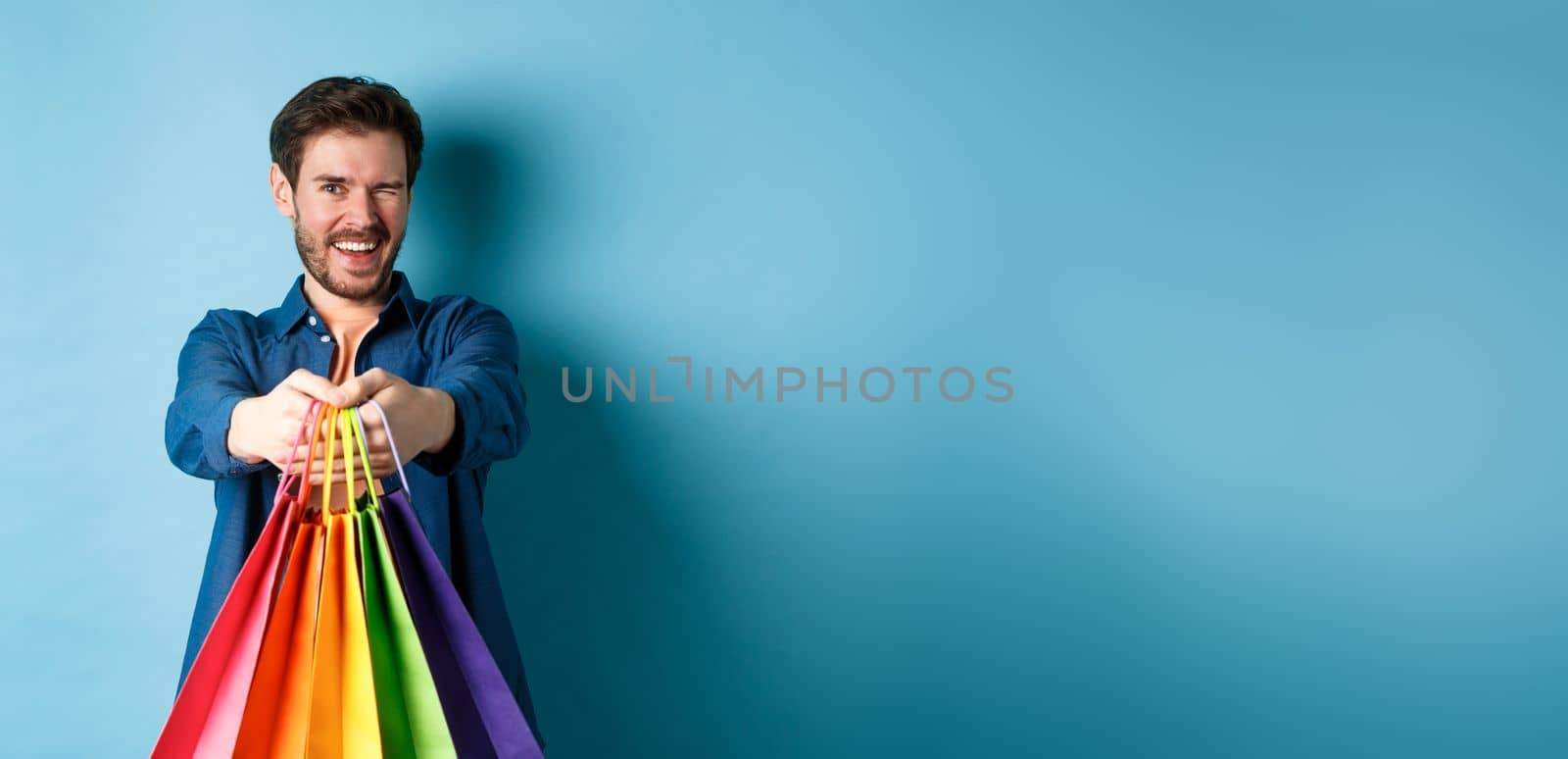 Cheerful handsome man winking and smiling, extending hands with colorful shopping bags, standing on blue background.