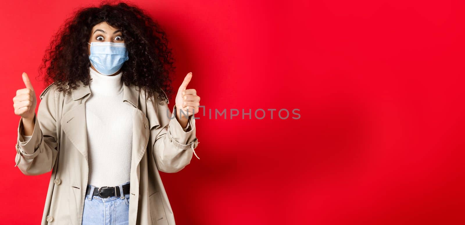 Covid-19, pandemic and quarantine concept. Excited girl with curly hair, wearing trench coat and medical mask, showing thumbs up in approval, red background.