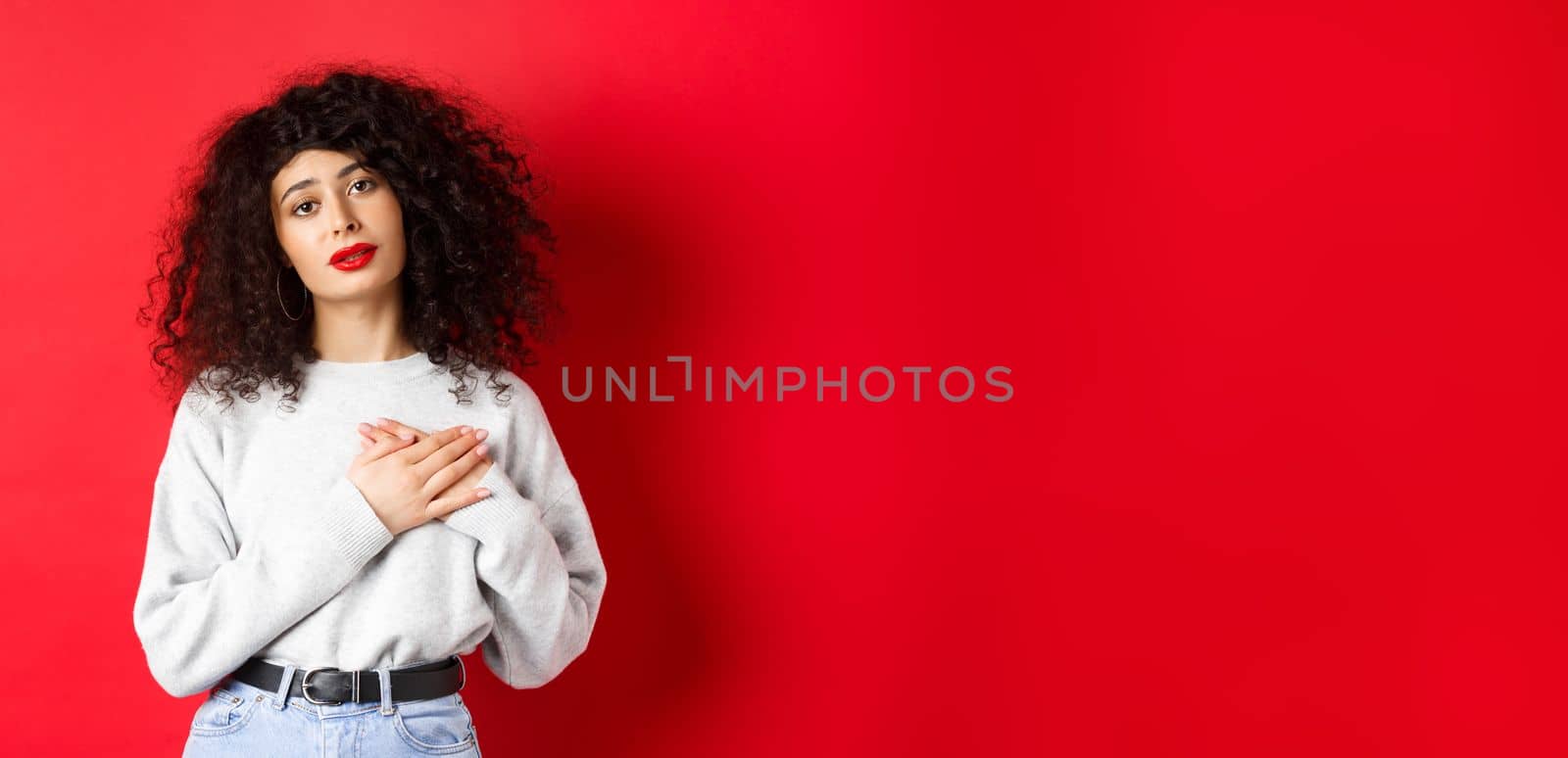 Beautiful woman with curly hair feeling touched and thankful, holding hands on heart and looking with affection at camera, red background by Benzoix