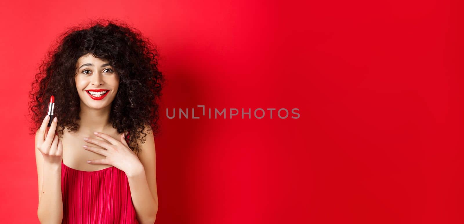 Beauty and make up concept. Beautiful female model with curly hair, wearing evening dress, showing red lipstick and smiling, standing on white background by Benzoix
