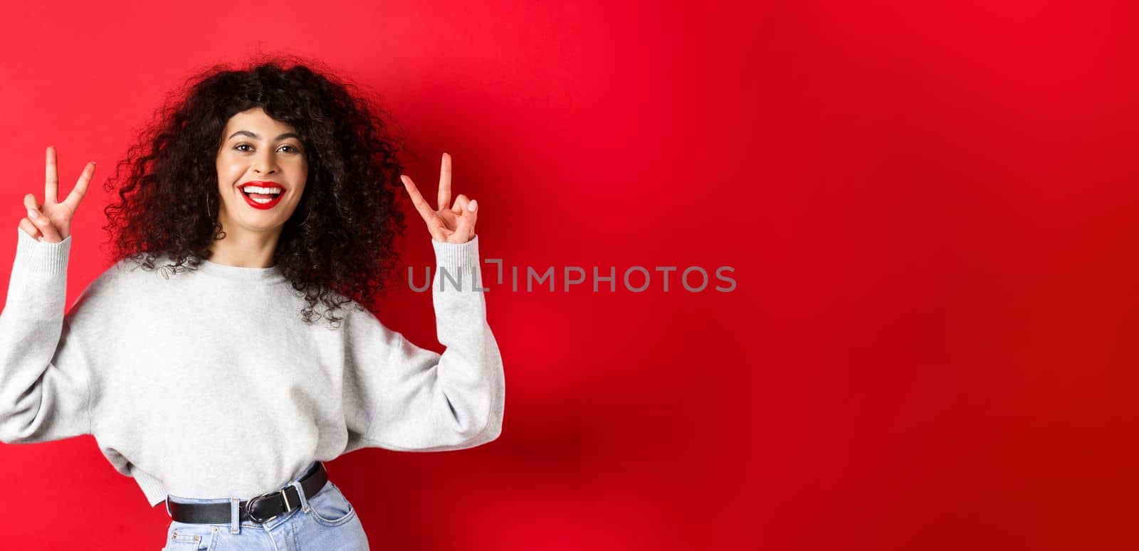 Stylish young woman with curly hairstyle, smiling happy and showing peace signs, standing in casual sweatshirt on red background by Benzoix