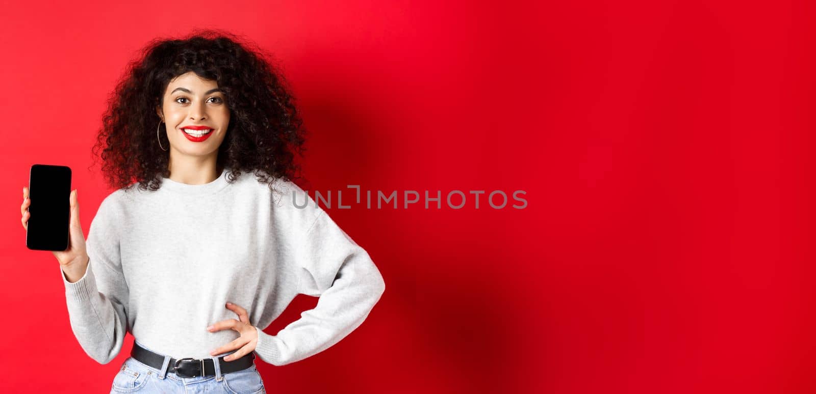 Attractive smiling woman showing empty smartphone screen and looking happy, advertising online store or application, standing against red background.