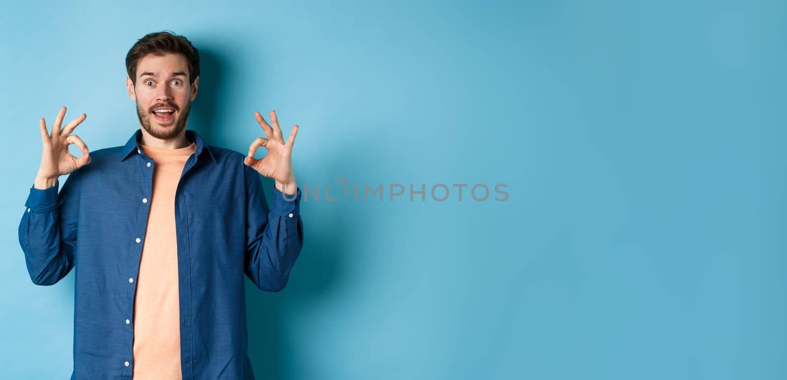 Impressed man praising something awesome, showing okay signs and looking amazed, complimenting good thing, standing on blue background.