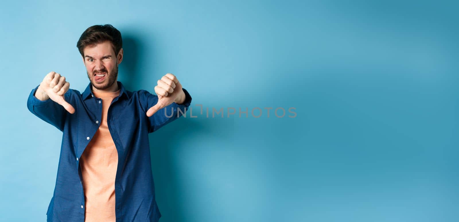 Disgusted guy express negative opinion, showing thumbs down and tongue, frowning upset, disapprove something bad, standing on blue background.