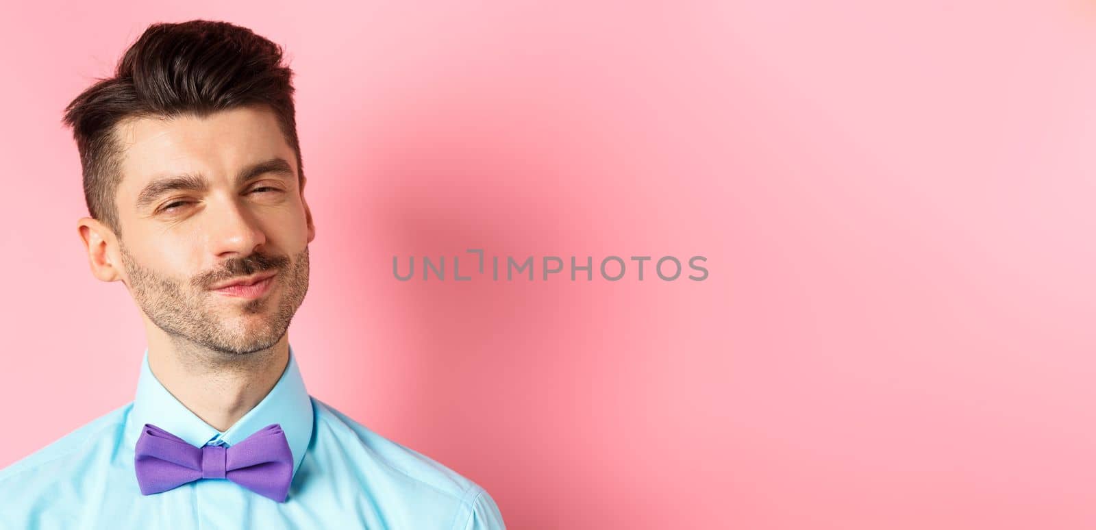 Close-up of smiling young guy with moustache, squinting intrigued, listening to something interesting, standing in bow-tie on pink background.