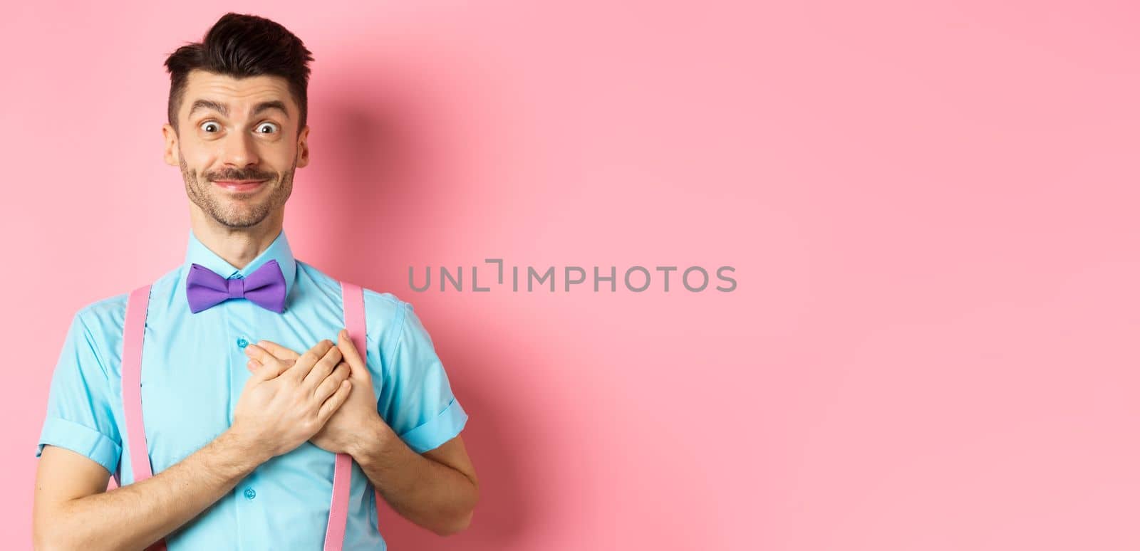 Cheerful young man with moustache, wearing shirt and bow-tie, holding hands on heart and smiling grateful, saying thank you, standing on pink background by Benzoix