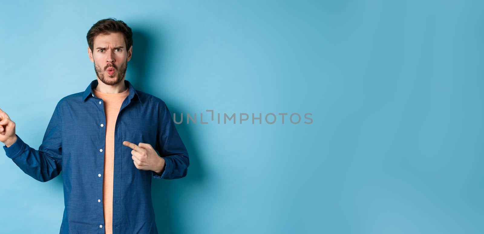 Shocked and offended young man frowning and looking hurt, pointing fingers left at empty space, complaining on something, standing on blue background.