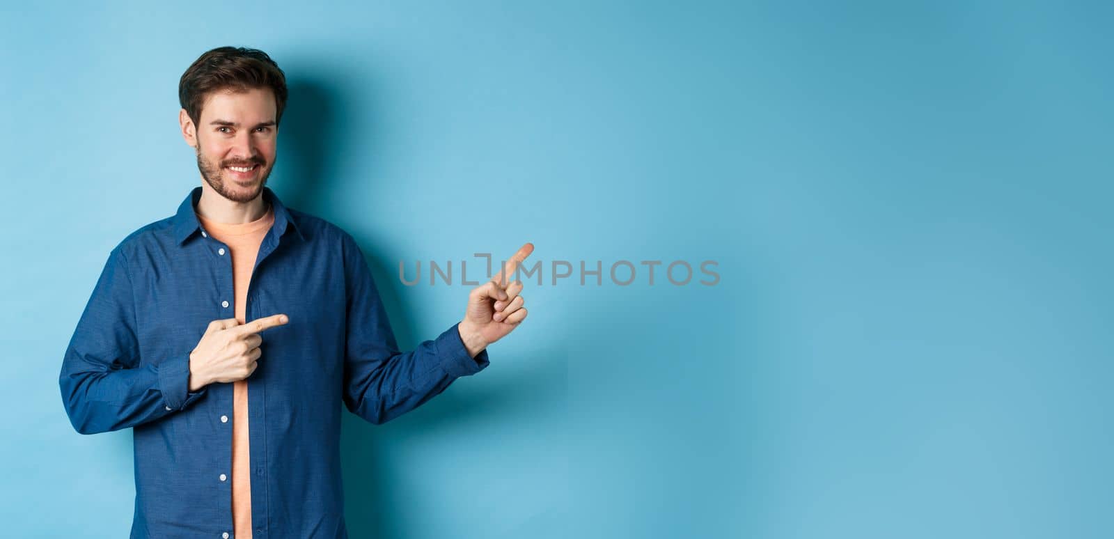 Smiling confident man showing logo, pointing fingers right at empty space and looking at camera, standing on blue background.