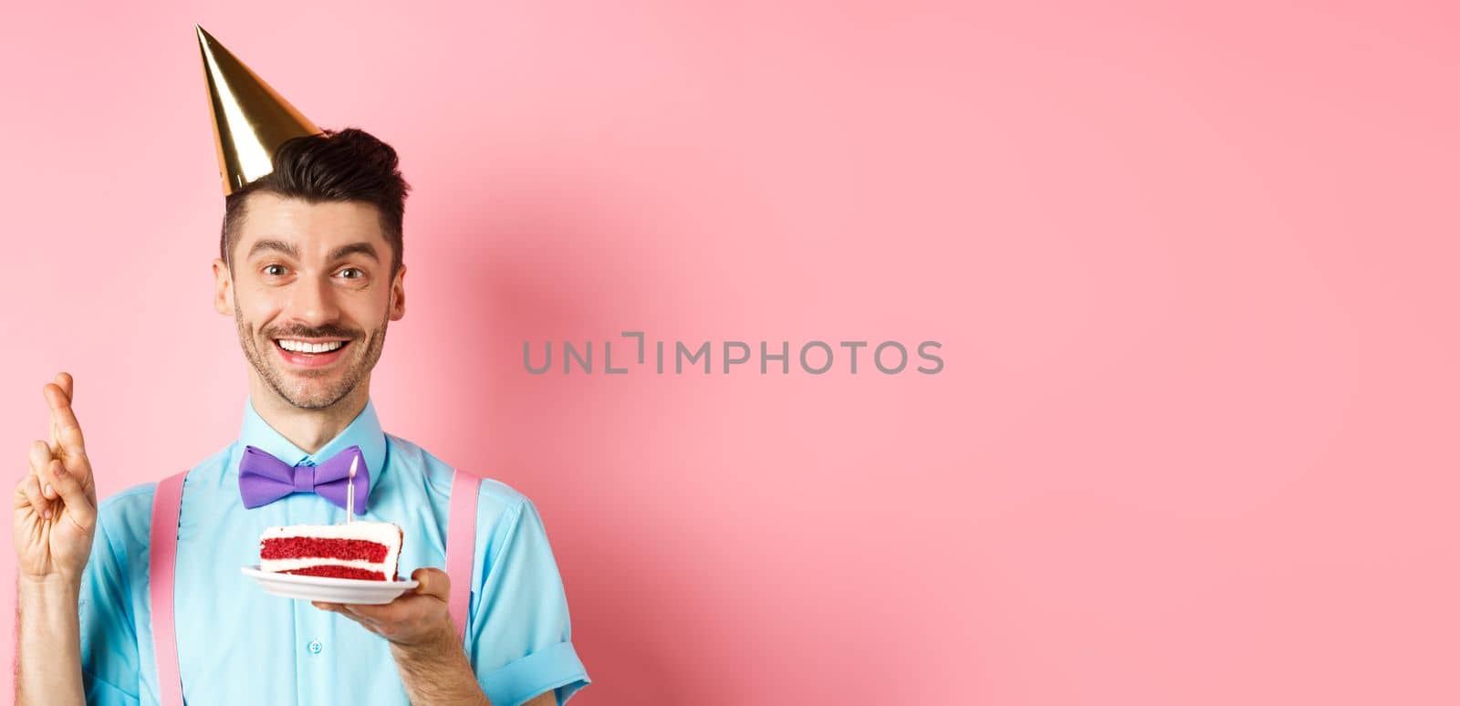 Holidays and celebration concept. Happy young man enjoying birthday party, wearing cone hat and cross fingers, making wish on bday cake with candle, pink background by Benzoix