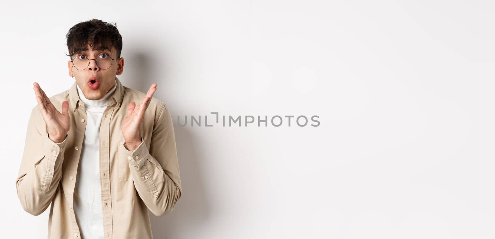 Portrait of surprised hipster guy in glasses checking out awesome promo, say wow and gasping amazed, raising hands up with disbelief, standing on white background.