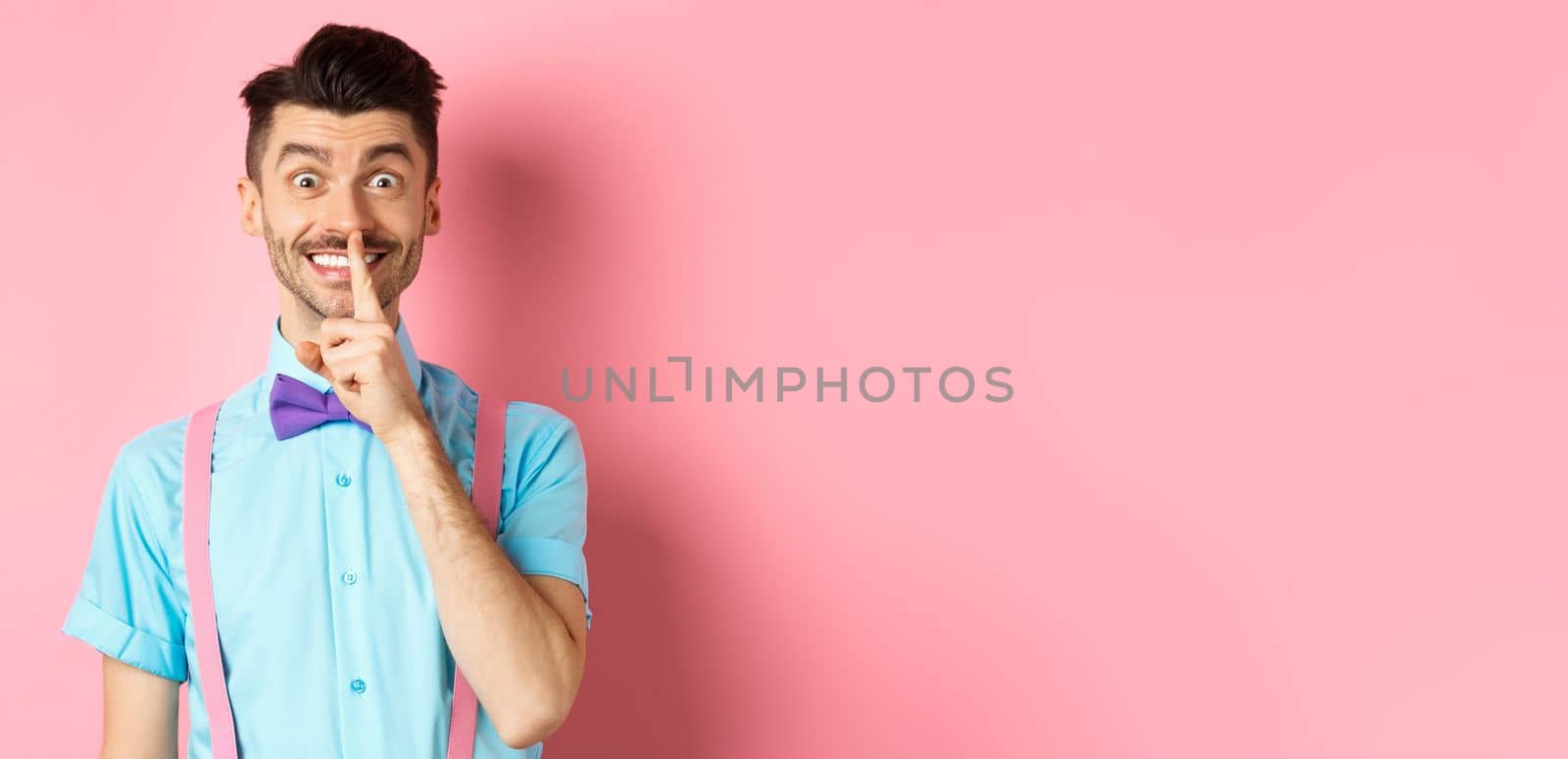 Cheerful young man making surprise, shushing at camera with happy smile, asking to keep voice down, be quiet, standing on pink background.