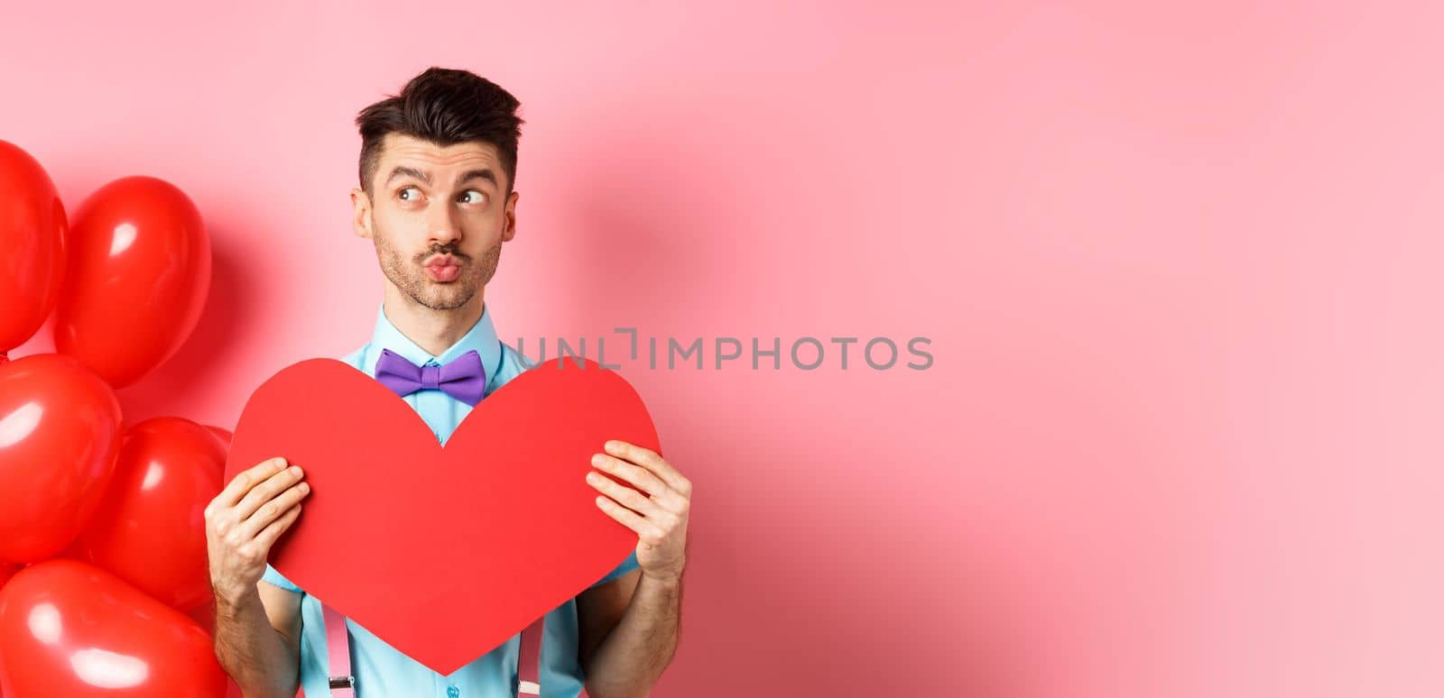 Valentines day concept. Charming young man in bowtie waiting for soulmate with big red heart cutout, looking left and dream of love, pink background.